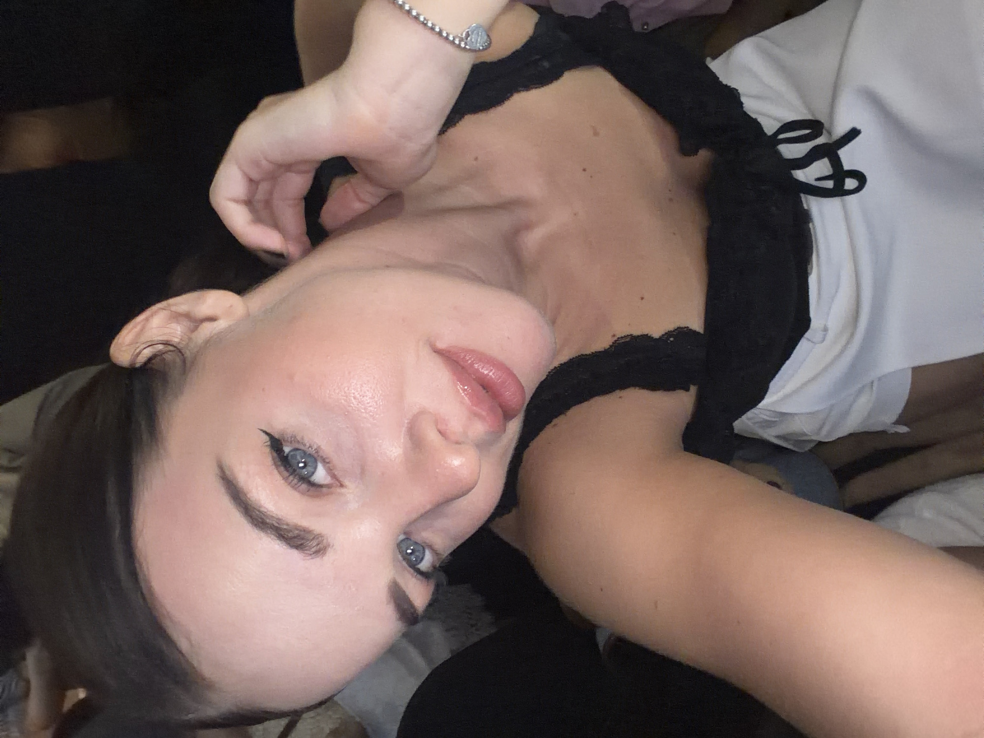 A young adult female is centered in a close-up, slightly angled portrait shot. She possesses dark brown hair, pulled back, and light skin. Her blue eyes are accentuated with black winged eyeliner, and her eyebrows are well-defined. Her lips are full and coated with a light, glossy lipstick. She wears a black lace-trimmed camisole over a white garment featuring black ties at the front. A silver beaded bracelet with a heart-shaped charm adorns her left wrist. Her right hand is raised, with fingers gently touching her neck area behind her right ear. The background is dark and blurred, suggesting an indoor, low-light environment with other indiscernible presences. The scene is located in Nha Trang Ward, Vietnam.