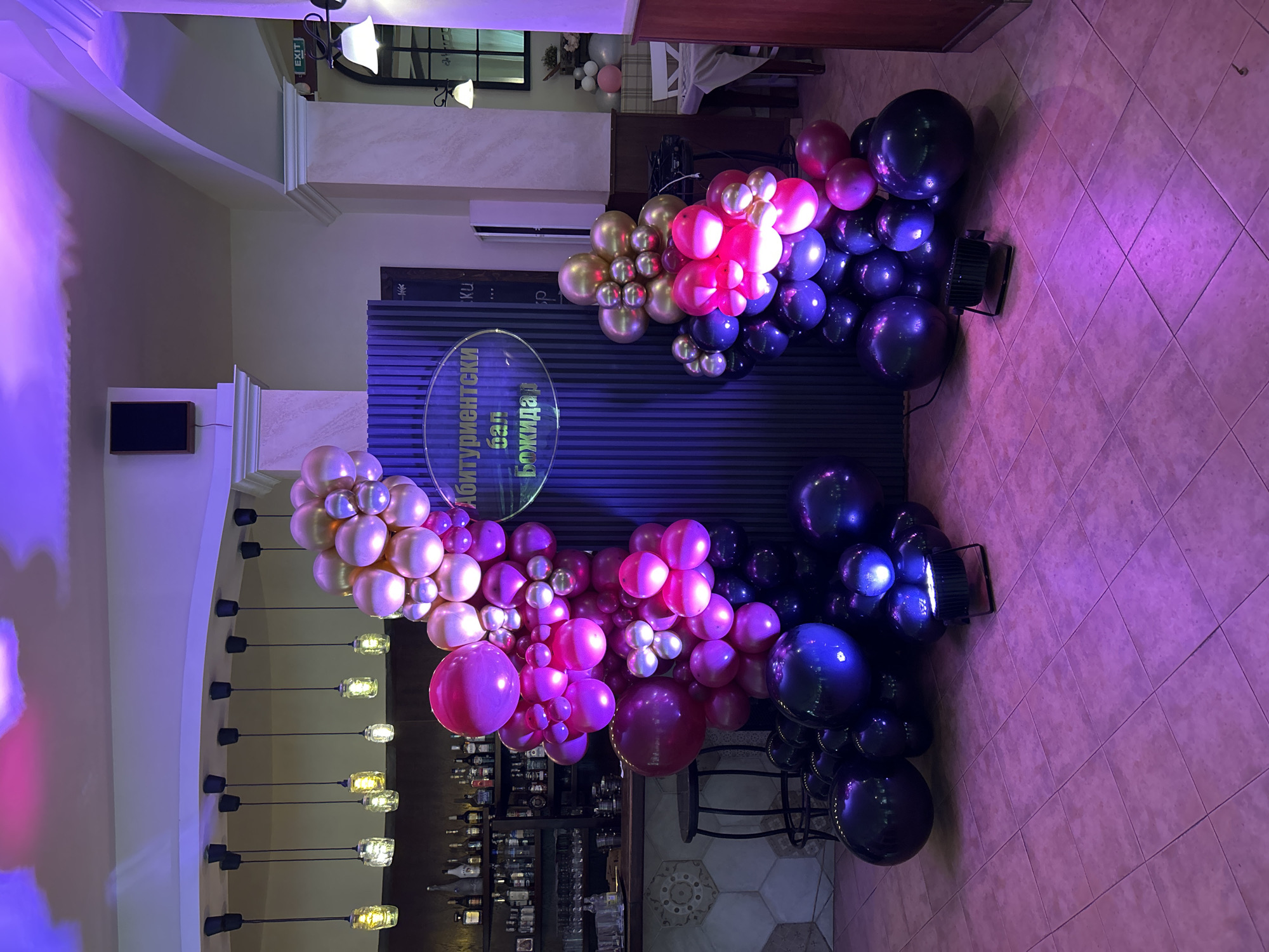 The image shows a decorated event space, likely a restaurant or bar, in Plovdiv, Bulgaria. Two balloon arches flank a backdrop sign written in Bulgarian, possibly for a graduation celebration. The arches are composed of metallic gold, rose gold, magenta, and black balloons. A lighting fixture with exposed bulbs hangs from the ceiling on the left. The floor is tiled in a beige geometric pattern. Two stage lights are situated on the floor next to the balloon arches. In the background, the bar is visible, with bottles of alcohol arranged on shelves. Overhead is an "exit" sign. The space is illuminated with purple lighting.
