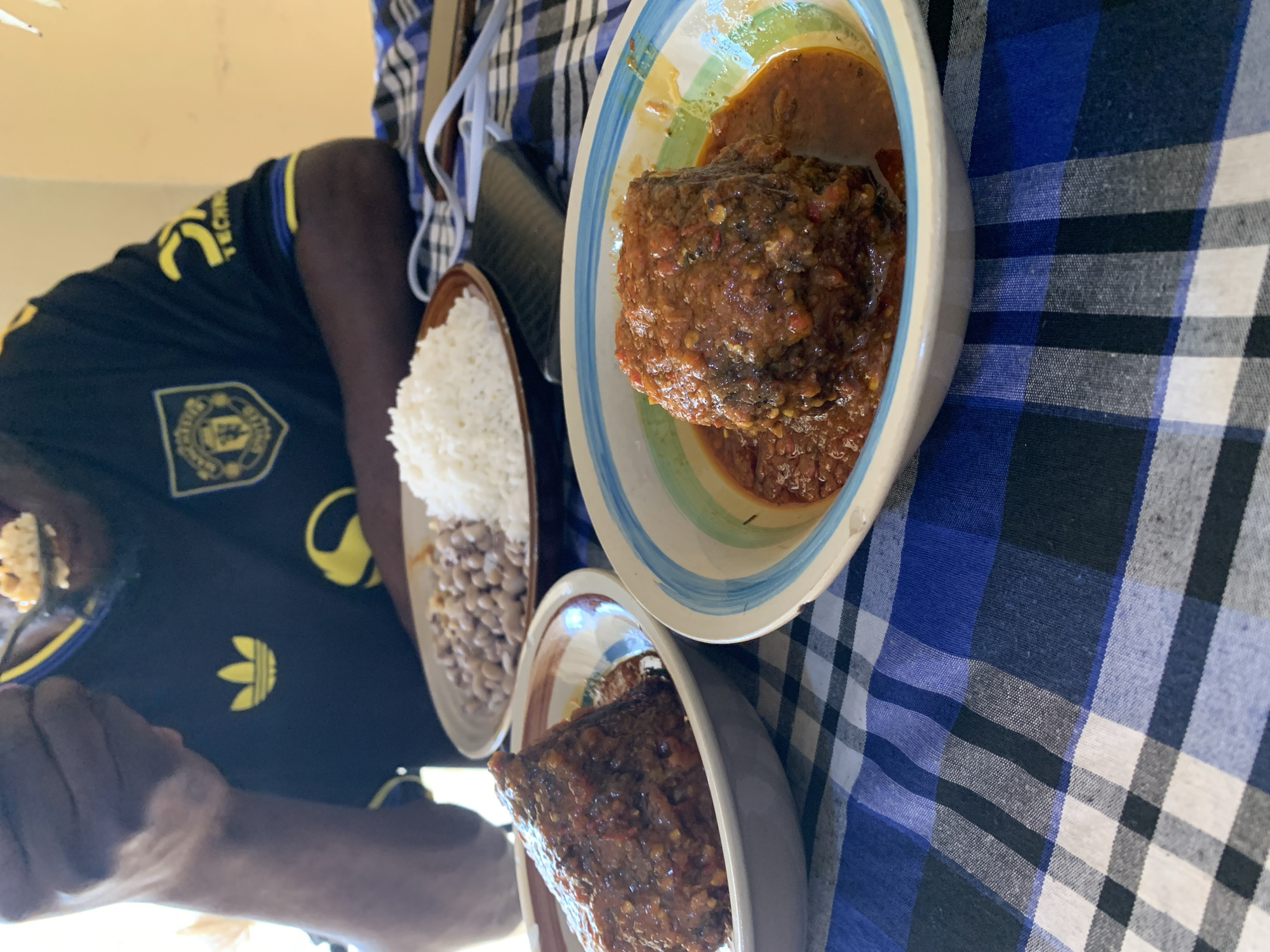 This image, captured in New Stadium, Nigeria, depicts an individual seated at a table, actively consuming a meal. The person, identifiable by their dark-skinned arm and hand, is seen bringing a spoon filled with light-colored food to their mouth. They are wearing a black short-sleeved t-shirt featuring a yellow Adidas logo on the chest, a Manchester United crest on the left breast, and a yellow "TECNO" logo on the right sleeve.
The table is covered with a blue, white, and black plaid tablecloth. Three food items are visible on the table. In the background, a brown plate contains a serving of white rice alongside light brown beans. In the foreground and mid-ground, two ceramic bowls hold a dark, solid food item generously coated in a thick, reddish-orange sauce, likely indicating a stew or curry. The bowl closer to the viewer has distinct blue and green stripes on its rim, while the other is a plainer beige with a brown rim. Also present on the tablecloth are a dark wallet or phone case and white cables, possibly belonging to headphones or a charging device. The background features a light-colored wall, with a soft light source emanating from the left side of the frame.