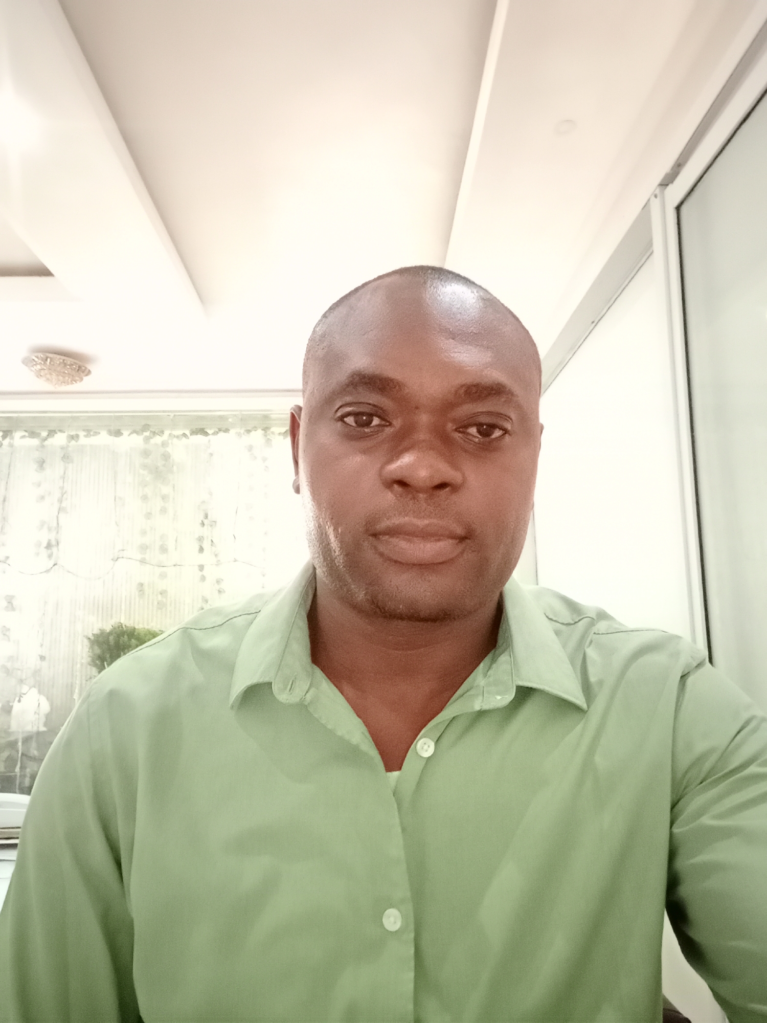 A medium shot captures a man with dark skin, bald head, and a green button-down shirt. He is centered in the frame and appears to be indoors. Behind him, a window is partially visible, revealing a blurred background that hints at an outdoor environment with vegetation. The indoor setting suggests a room with a modern ceiling design and a light fixture visible at the top of the frame. The man's expression is neutral as he looks directly at the camera. There is no evidence in the image to confirm the location in Gwarinpa, Nigeria.