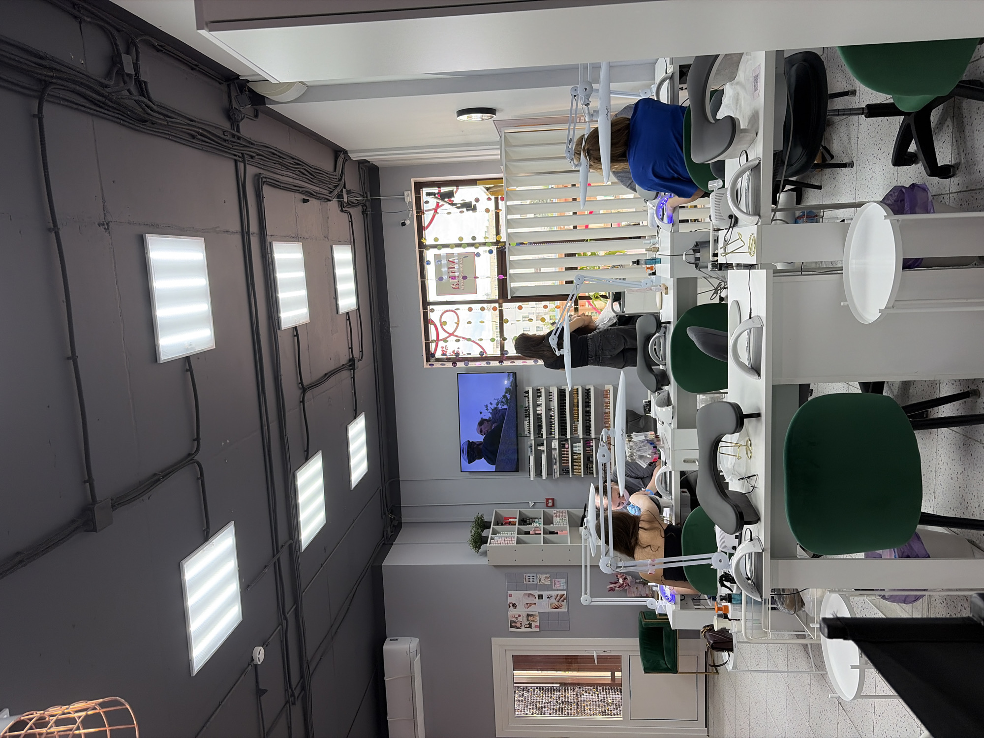 The image depicts the interior of a nail salon in Imeni Morozova, Russia. The scene is brightly lit by fluorescent ceiling lights, with additional task lighting at the workstations. Several women are present, appearing to be technicians and clients, engaged in nail services. One woman in a blue top is working on another's nails, with a UV lamp visible on the table. Another woman, wearing a black top and jeans, is seated at a workstation, seemingly receiving a nail treatment.
The salon features multiple white workstations with ample storage for nail polish and supplies. Green and gray chairs are placed around the workstations. A television screen mounted on the wall displays an image of two people. A window with blinds is visible, offering a glimpse of the outdoors, though the time of day or weather are not discernible. Various objects, including bottles, brushes, and other beauty tools, are neatly arranged on the tables. The overall impression is one of a professional and busy beauty establishment. There is no visible text that is legible.