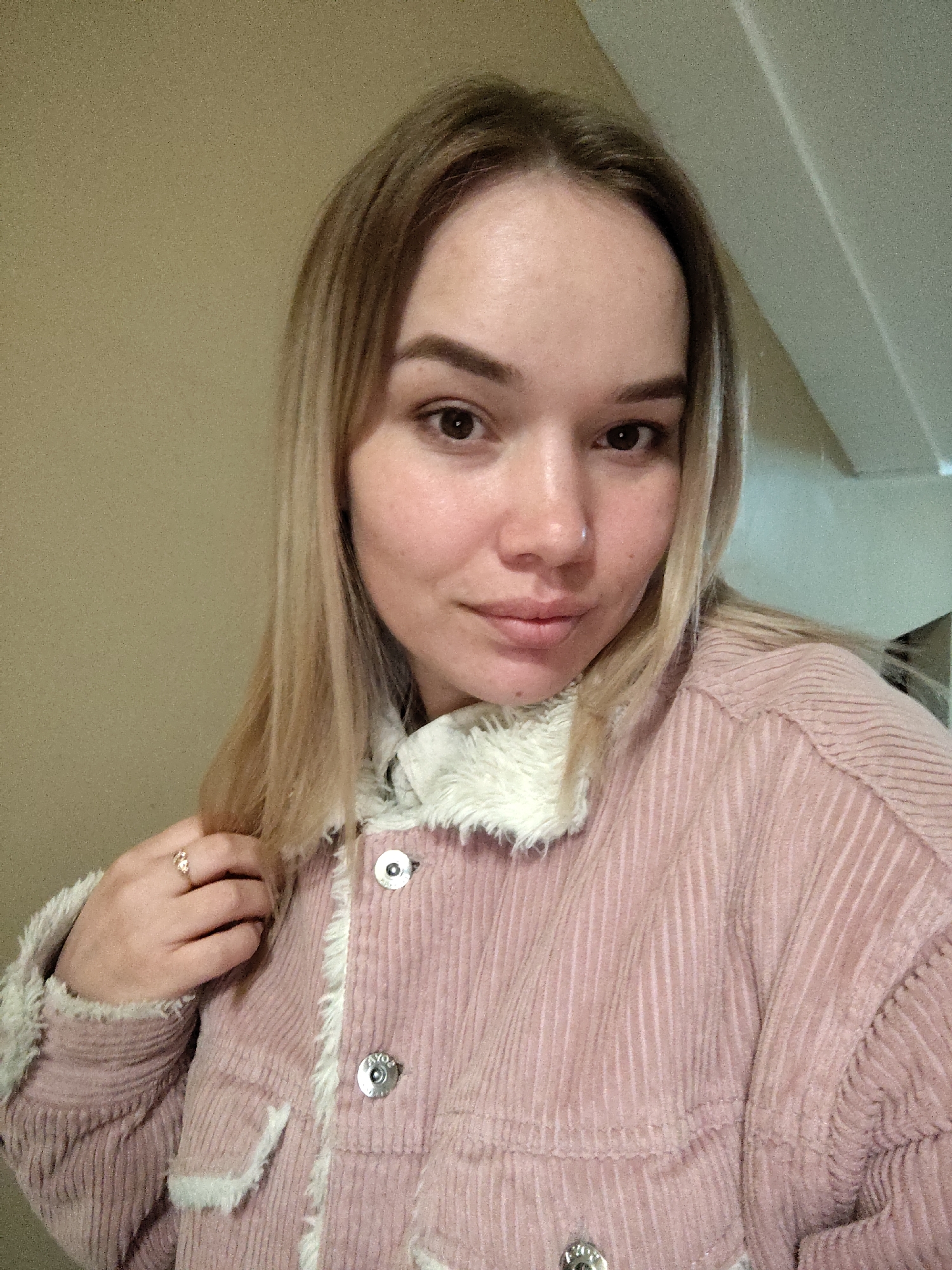 A young woman with light brown hair and dark eyes is depicted in a close-up, indoor portrait. She gazes directly at the camera with a calm, gentle expression and a subtle smile. She wears a pink corduroy jacket featuring prominent vertical ribs and white fluffy trim along the collar and cuffs. A small silver stud nose piercing is visible on her left nostril. On the ring finger of her right hand, she wears a golden ring with a floral or cluster design, and her hand is gently touching a strand of her hair near her shoulder. The background is a plain, light beige wall, with a hint of a ceiling line or architectural detail in the upper right corner, indicating an indoor setting. One of the silver buttons on her jacket has faintly visible text, possibly reading "AYOS" or "AVOS". The scene suggests a relaxed atmosphere, likely in Марковский, Russia.