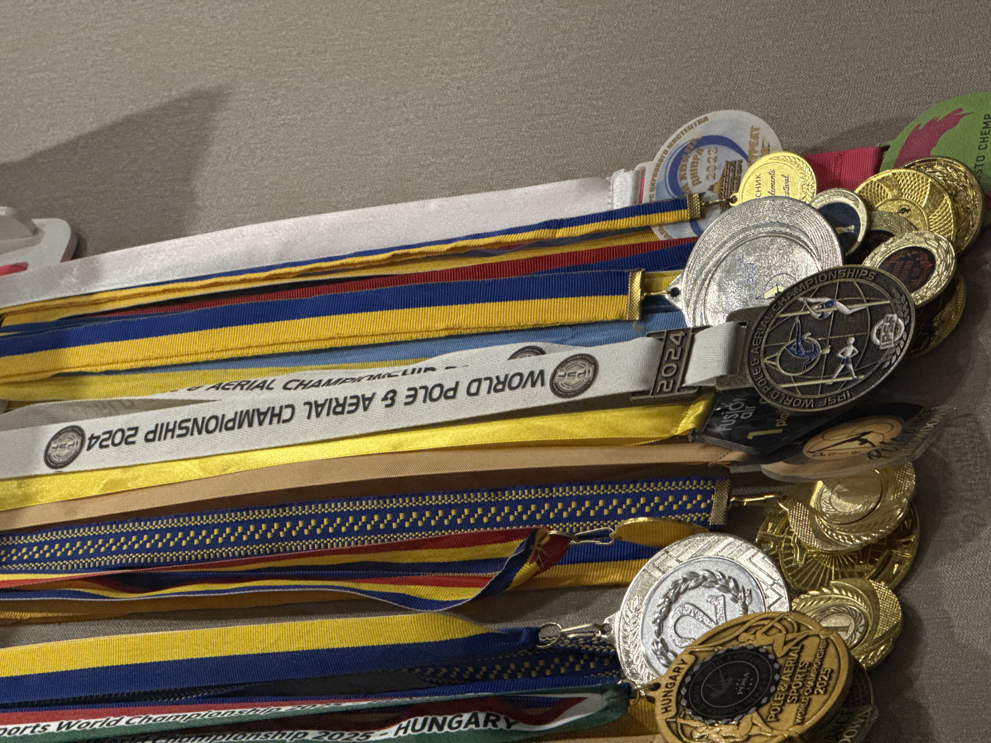 This media file displays a collection of medals and their accompanying ribbons, laid out on a textured surface, likely a table or floor. The medals vary in size, color (gold, silver, and bronze tones are visible), and design, with some featuring intricate engravings and logos. Several ribbons are multi-colored, displaying stripes in shades of blue, yellow, red, and white, while others are solid white or gold.
Visible text on the ribbons and medals indicates several events and organizations. Phrases like "WORLD POLE & AERIAL CHAMPIONSHIP 2024," "SPORTS WORLD CHAMPIONSHIP 2025 HUNGARY," and "IPSE" are clearly legible. Additionally, Cyrillic text appears on some medals, including "ЦИРКОВОГО МИСТЕЦТВА ДНІПРА 2023," which translates to "CIRCUS ARTS OF DNIPRO 2023," suggesting a connection to an event in Dnipro, Ukraine. Some medals also show the word "HUNGARY" and "POSA."
The arrangement of the medals suggests they are awards for achievements in pole and aerial sports. The overall scene is that of a display of accomplishments, likely personal or for a competition. There are no people or specific environmental cues like time of day or weather, as the focus is entirely on the objects and their inscribed details. The lighting is even, suggesting it might be an indoor setting.