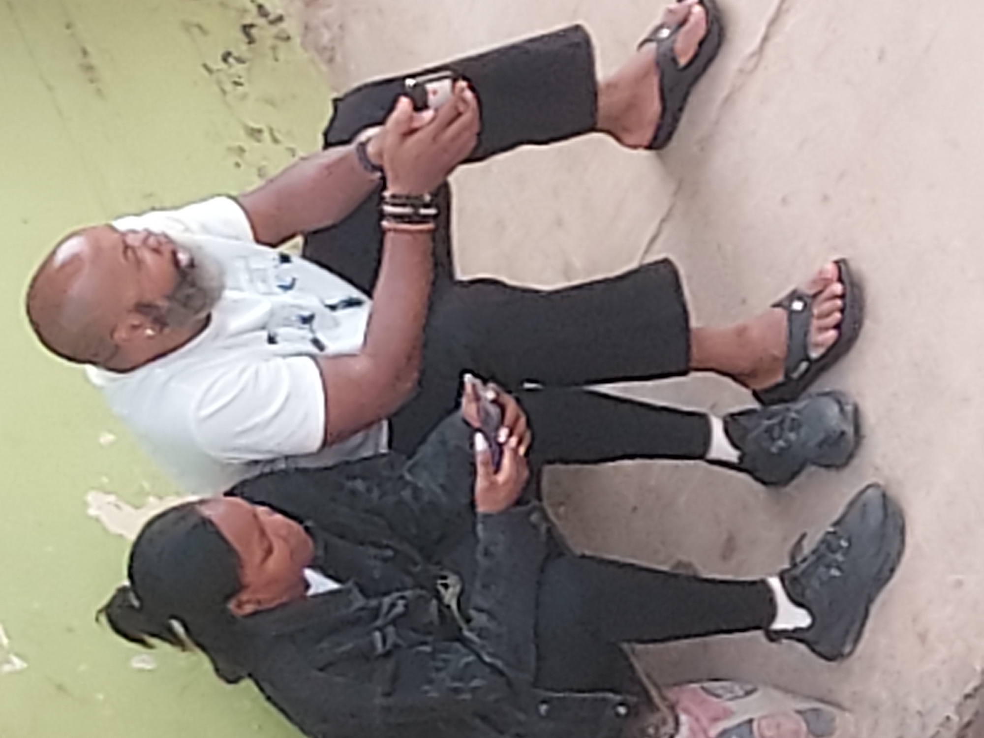 The image, rotated 90 degrees clockwise, shows two individuals, a man and a woman, seated on a light-colored ground surface, possibly concrete or pavement. A green patch is visible in the background to their left. The scene appears to be an informal, possibly outdoor or semi-open setting, likely in Jos, Nigeria.
The man, positioned further back, appears middle-aged with a bald head and a gray beard. He is wearing a white short-sleeved t-shirt, black trousers, and black flip-flops. A gold stud earring is visible in his left ear, and he has several beaded bracelets on his left wrist. He holds a white smartphone horizontally in both hands, looking intently at its screen.
The woman is seated closer to the foreground, in front of the man. She has dark hair, which appears to be tied back. She is dressed in a dark, possibly black, long-sleeved jacket or shirt, black trousers, white socks, and dark sneakers. Like the man, she is holding a dark smartphone in her hands and is focused on its display. A multi-colored bag is partially visible on the ground beside her feet.
Both individuals are casually dressed and engrossed in their mobile phones, suggesting they are either resting, waiting, or simply passing time. There is no visible text on their clothing, phones, or in the surrounding environment. The overall mood appears calm and focused on their individual devices.