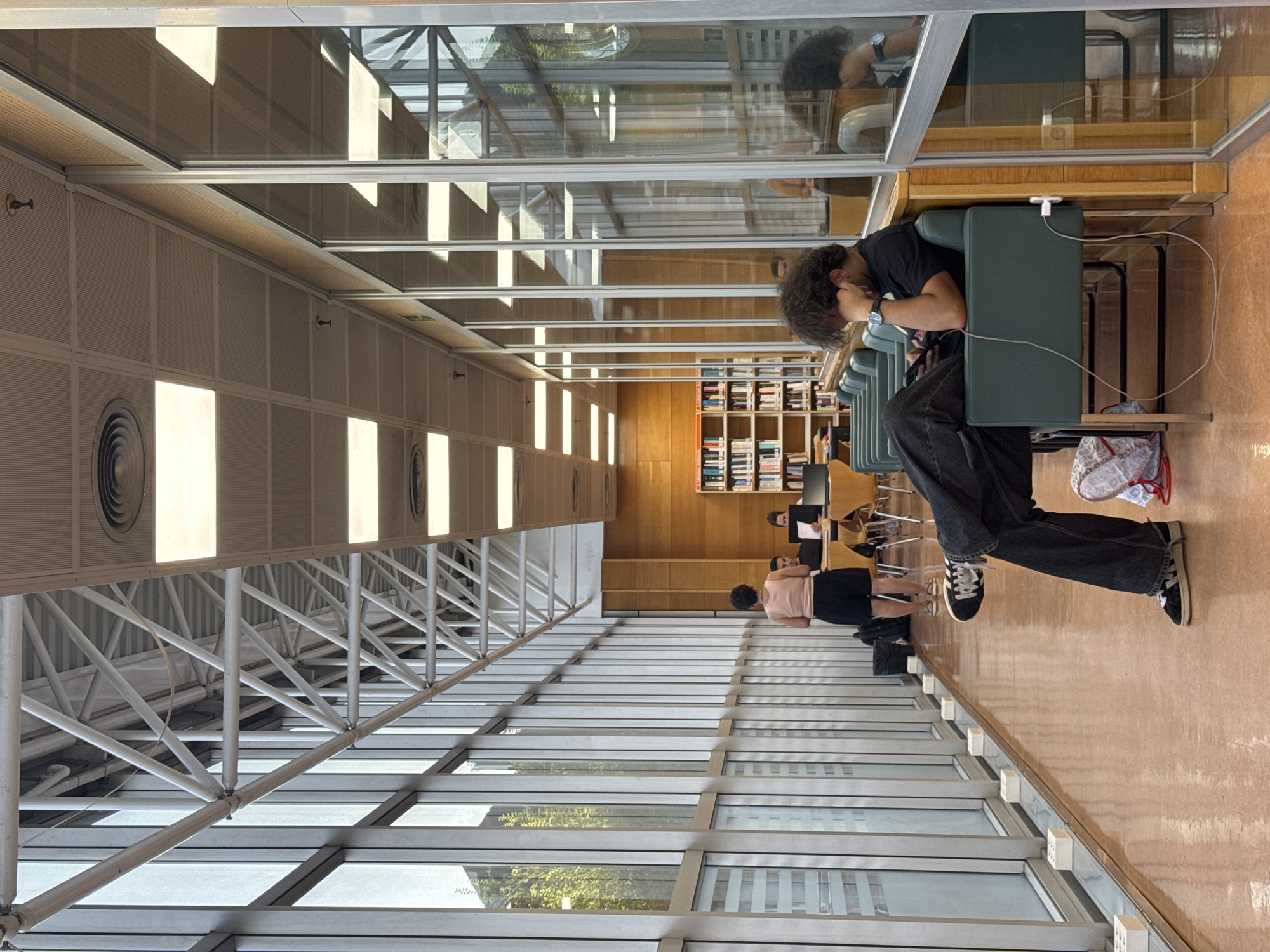 The image shows the interior of a modern library with large windows and a high ceiling with exposed beams. In the foreground, a person with curly hair is seated at a table, leaning their head into their hand. They are wearing a black t-shirt and dark jeans. A white charging cable is plugged into a device on the table and trails across the floor. Further back in the library, other individuals are visible, including someone in a pink t-shirt and black shorts and another person standing at a desk with a laptop. Bookshelves filled with books line the far wall of the library. The lighting suggests it is daytime, and the overall atmosphere appears calm and studious. The scene is set within a contemporary public building, likely a university or town library.