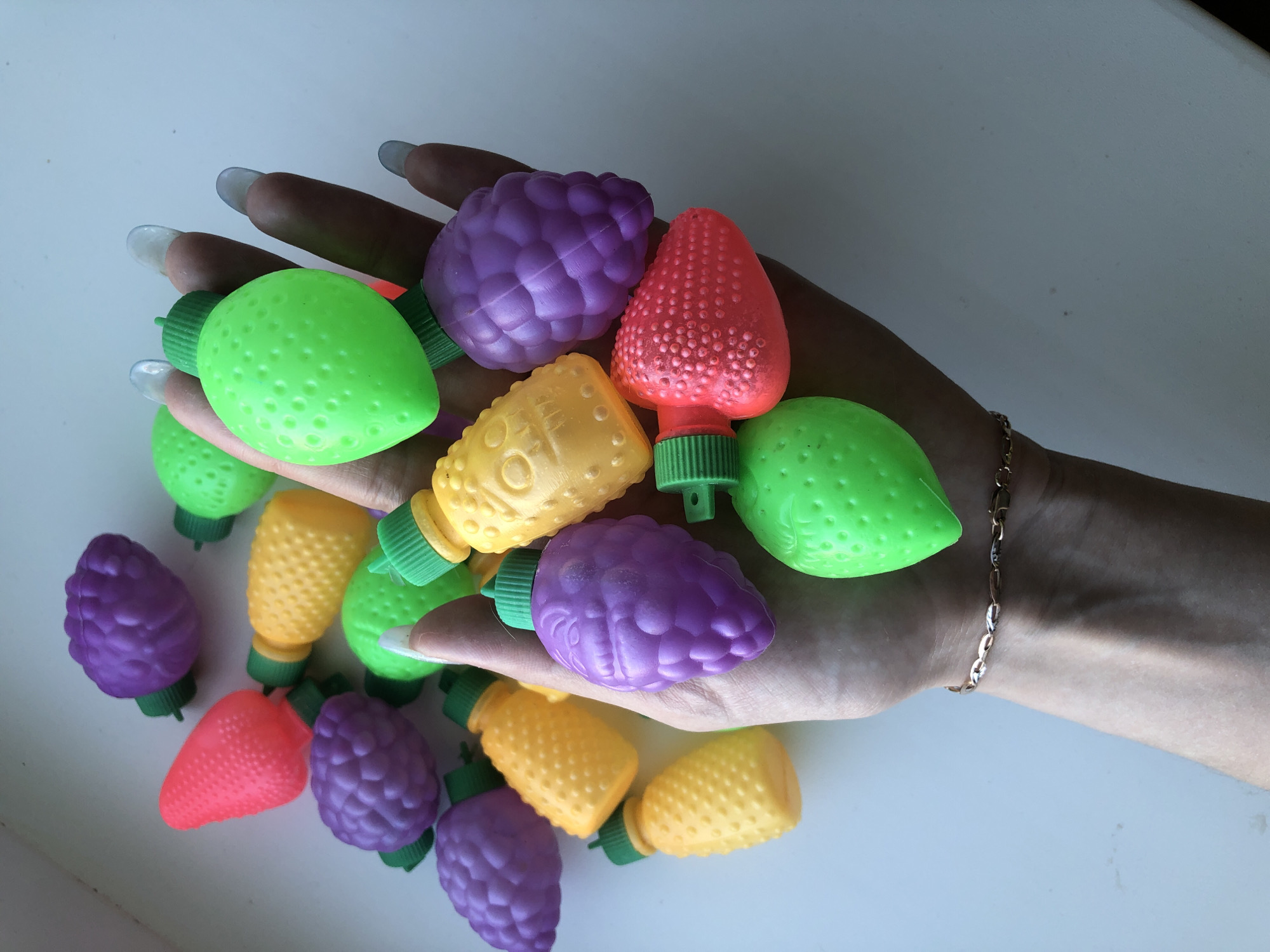 A light-skinned hand, adorned with elongated acrylic nails, holds an assortment of colorful plastic Christmas light covers shaped like fruits. The colors include green, purple, yellow, and red. The light covers are textured to resemble lemons, grapes, pineapples, and strawberries. A gold-colored chain bracelet is visible on the wrist. The background is a plain white surface. There is no indication of a location in the city of Noviy, Russia.