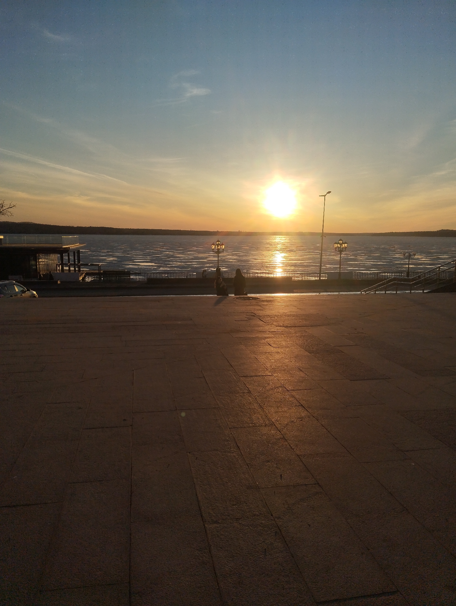 A wide-angle shot shows a paved square in Votkinsk, Russia, overlooking a body of water at sunset. The sky transitions from blue at the top to orange near the horizon, where the sun casts a bright reflection on the water's surface. The water appears to be partially frozen with streaks of ice. A few silhouetted figures are seated near the edge of the square, looking out at the water. Decorative lampposts and a modern street light line the embankment. A building with a balcony and a car are visible to the left. Stairs lead down to the embankment from the square on the right. The foreground is dominated by the paving stones of the square, which reflect the warm light of the setting sun. The overall impression is one of tranquility and beauty.