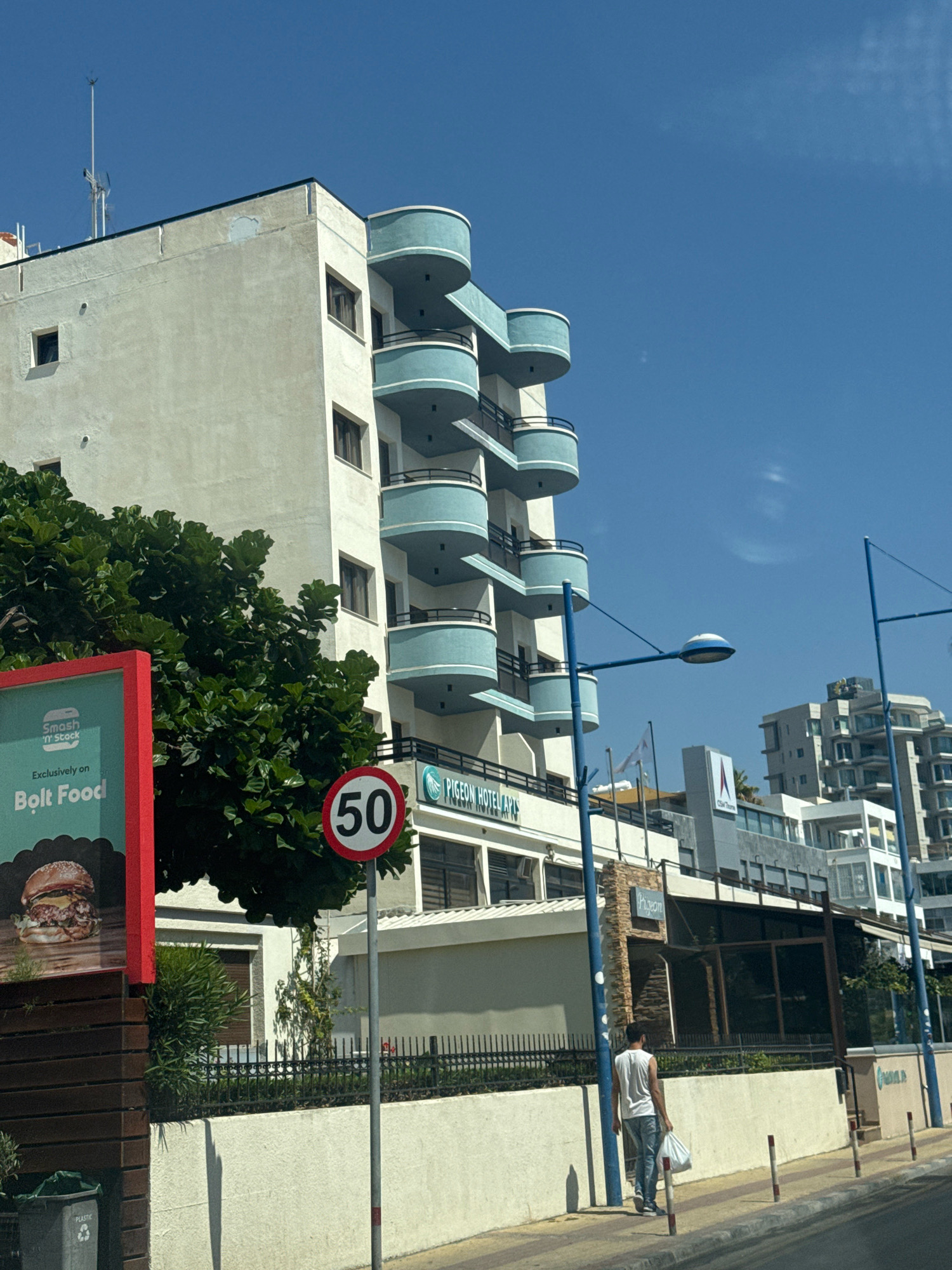 This is a full-shot, eye-level, outdoor photograph taken on a bright, sunny day in the Agios Tychona Community, Cyprus. The foreground features a dark brown wooden fence and a gray trash bin with "PLASTIC" written on it. A red and teal sign for "Smash 'n' Stack" and "Bolt Food" is visible on the left, displaying an image of a burger. To the right of the sign, a speed limit sign indicating "50" is mounted on a metal pole.
Behind the fence and sign, a white building with light blue balconies is the dominant structure, displaying architectural elements consistent with a hotel or apartment complex. The building has multiple stories with dark-framed windows. Next to this building, a sign reads "PIGEON HOTEL APTS".
Further in the background, other buildings of varying heights and architectural styles are visible, including a modern, multi-story building with white balconies and glass sections. A light blue street lamp with a curved fixture is positioned in front of the Pigeon Hotel.
A man wearing a white sleeveless shirt and blue jeans walks away from the camera on the sidewalk, carrying a white plastic bag. The sidewalk is lined with red and white bollards. The sky is clear and blue.