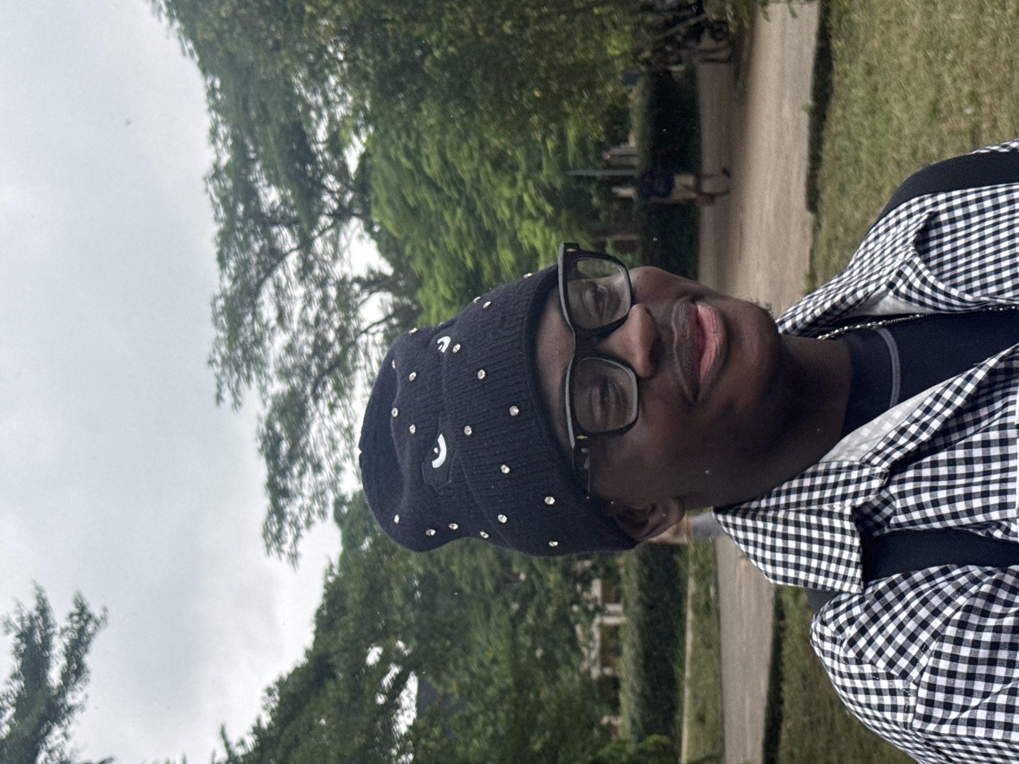 The image shows a close-up of a Black man standing outdoors. He is wearing a black beanie with a white eye design and rhinestones, black-rimmed glasses, and a black and white checkered shirt over a black t-shirt. A silver chain necklace is visible. He is smiling and looking slightly to his right. The background consists of lush green trees and foliage, with a path and some indistinct figures in the distance. The overall lighting suggests an overcast day. The setting appears to be a park or a well-maintained outdoor area. Given the provided context of Ihiagwa, Nigeria, this scene could be depicting a typical day in one of the city's green spaces or recreational areas.