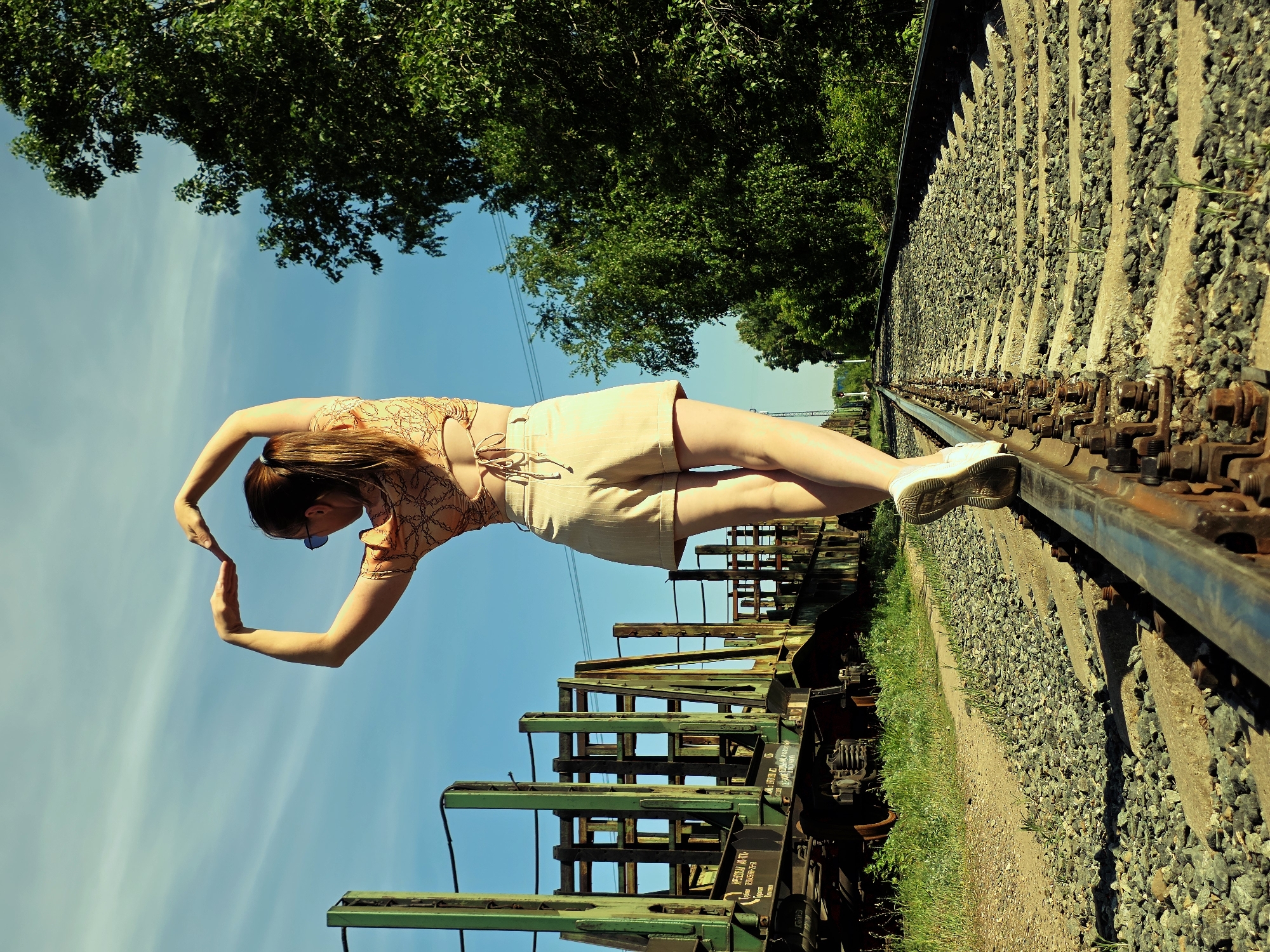 A young woman in sunglasses, a patterned orange top, and a beige skirt stands on a railroad track, her weight supported by her right leg and right foot resting on the rail. She balances, using her arms to create a heart-shaped form above her head. Her left leg is extended out over the railcar directly beneath her. Her right shoe is white, and the railcar is green and has an unidentifiable inscription written on it. The railroad tracks are made of metal and run alongside gravel. Green trees are overhead, and the sky is blue with wispy white clouds. The location is Chaikovsky, Russia.