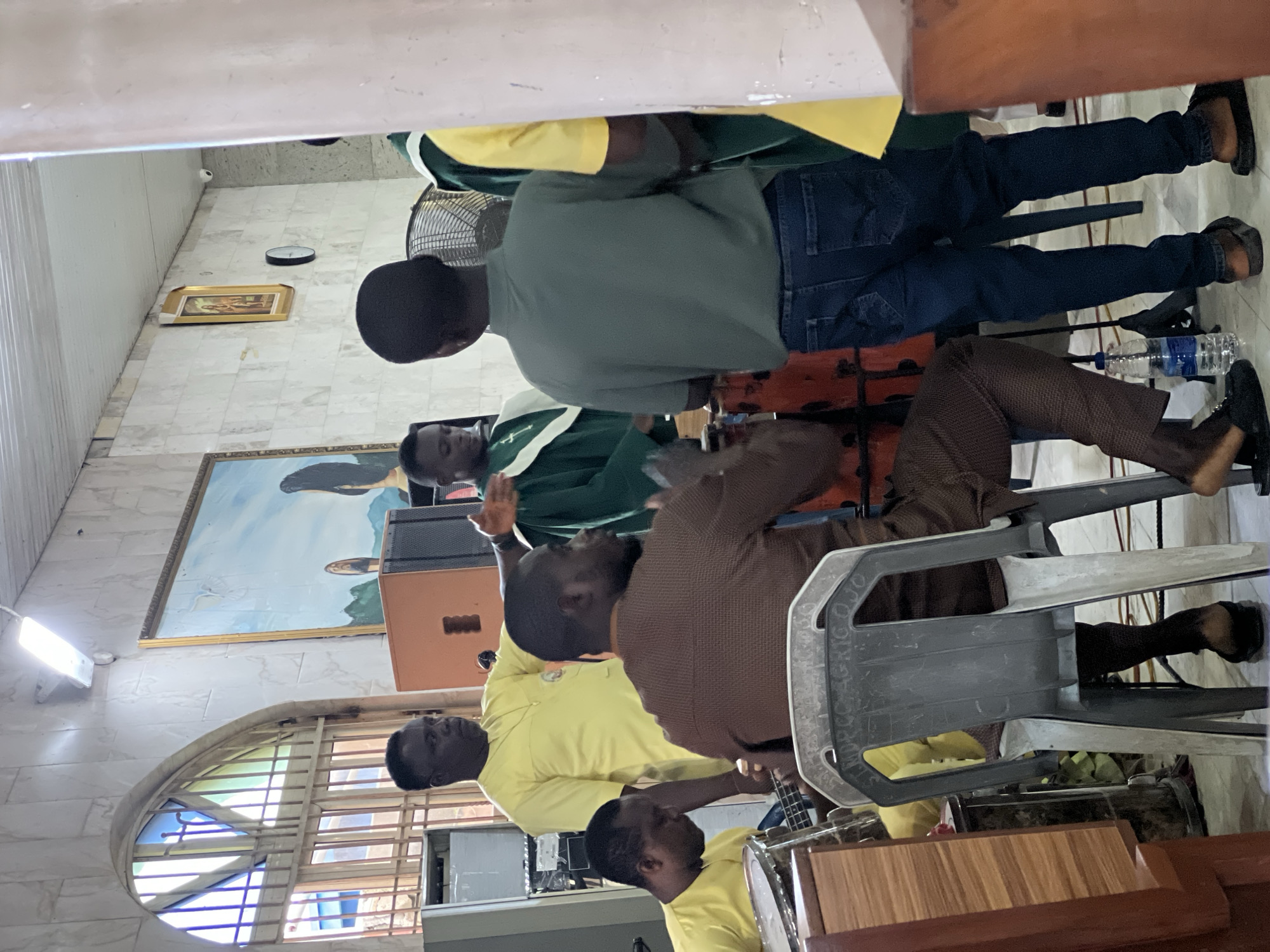 In Lagos, Nigeria, a group of individuals are gathered inside what appears to be a church. The environment is characterized by tiled walls and a painted mural on the wall, depicting a religious scene. In the foreground, a man in a brown patterned outfit is seated, facing left, with his right hand resting on a drum. To his left, another man in a yellow shirt is playing a bass guitar. Behind them, a man in a yellow shirt stands with his arms outstretched, seemingly directing. Further back, individuals in green robes are visible, one playing a drum. A large fan is situated behind the group, and a bottle of water is on the floor. The scene captures a moment of musical activity, possibly during a service or rehearsal.
