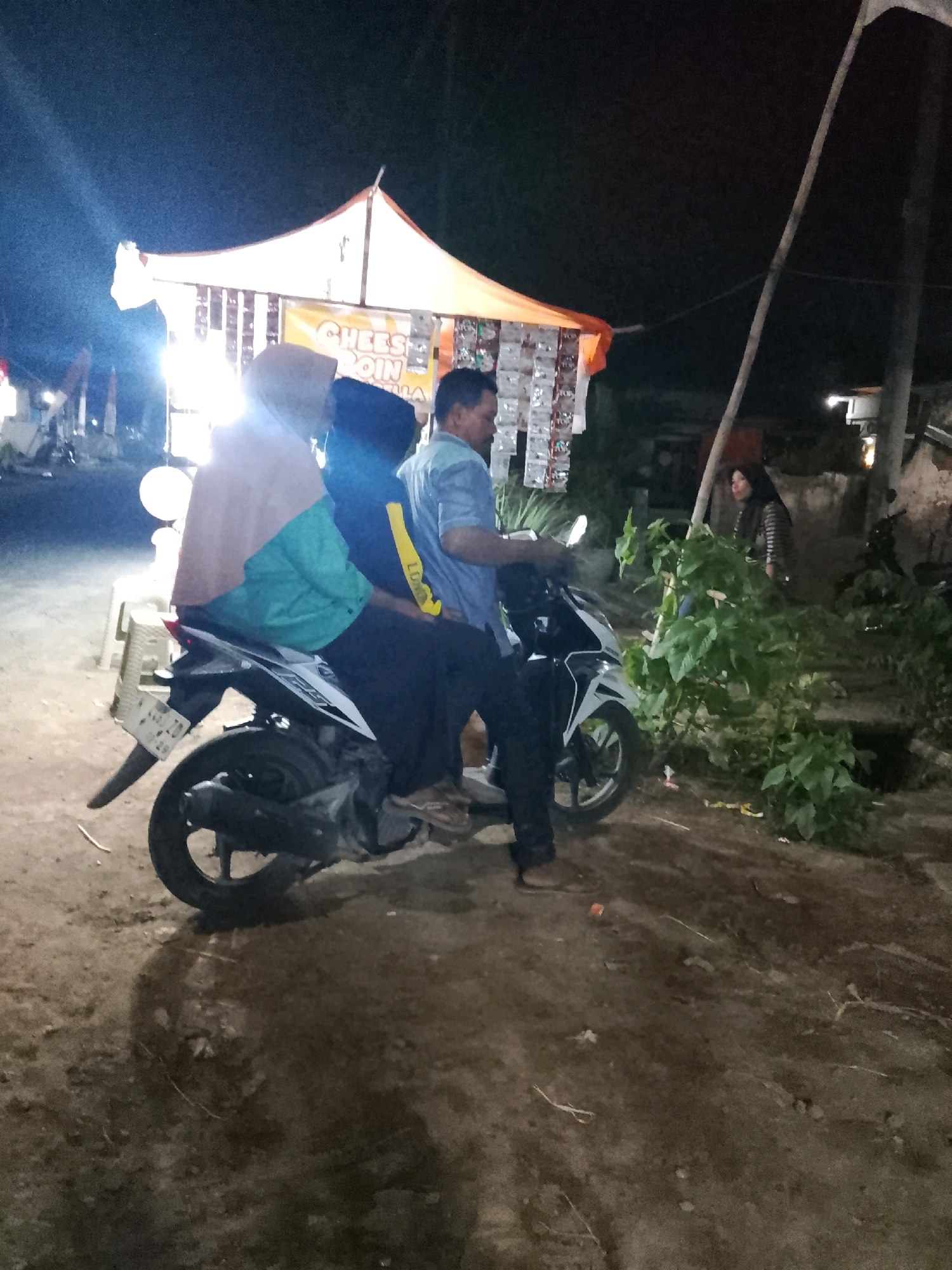 At night in Sukorejo, Indonesia, a motorcycle is parked on a dirt road in front of a stall. Three people are visible around the motorcycle. A man in a light blue shirt is seated on the motorcycle, and two individuals, possibly women, are behind him, also on the motorcycle. One of them is wearing a headscarf that is light pink and turquoise, and the other is wearing a dark top with a yellow stripe down the back. A woman in a striped shirt is standing further away, near some greenery and a pole. The stall appears to be illuminated, with a banner that reads "CHEES" and "QIN" visible. The ground is uneven and appears to be composed of dirt and some debris. The overall lighting suggests it is nighttime, with artificial lights from the stall and possibly other ambient light sources.