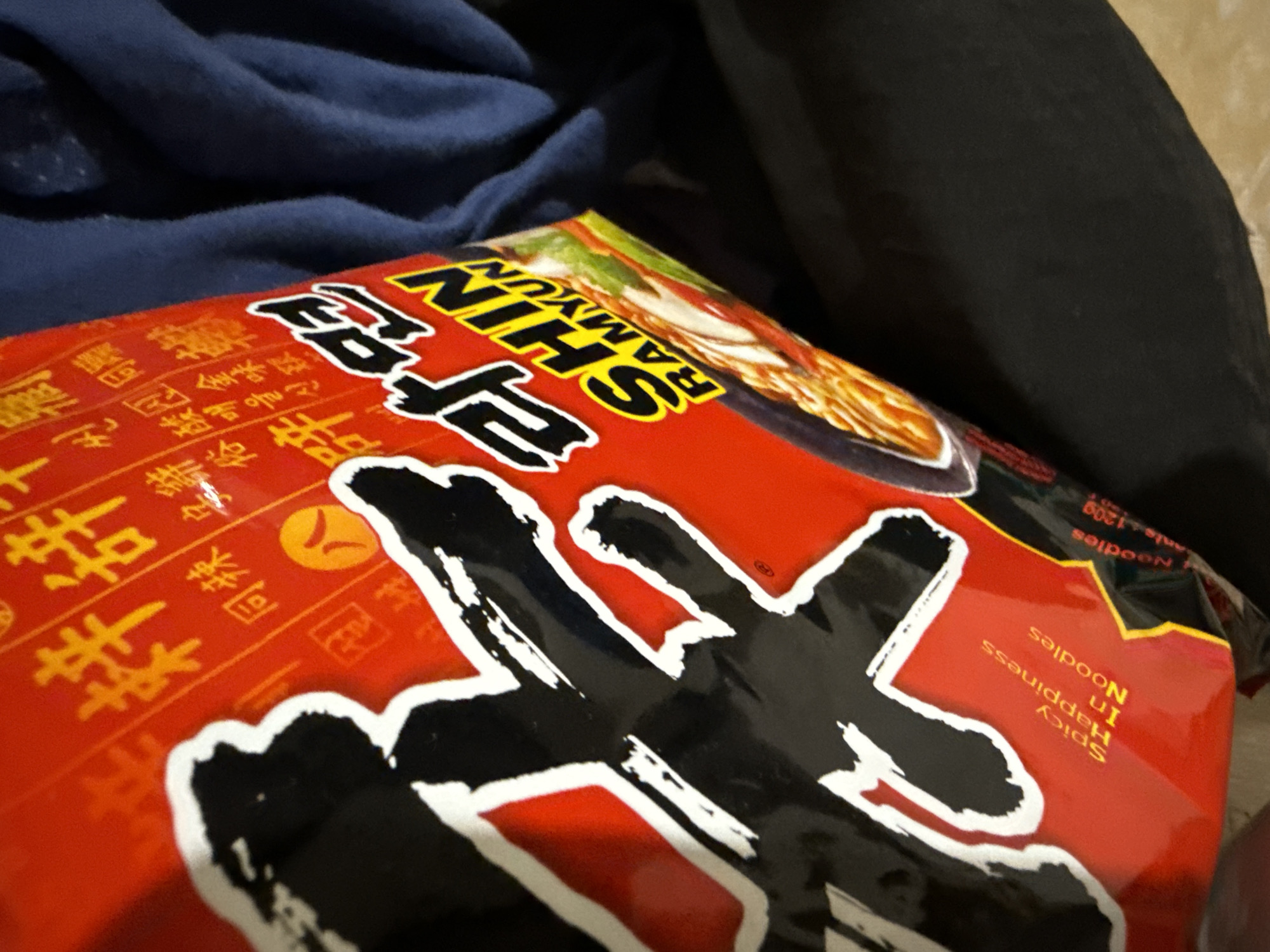 The image shows a close-up of a red package of Shin Ramyun instant noodles. The package is partially obscured by dark fabric, likely clothing, and rests on a blue surface, possibly a textile. The Shin Ramyun logo is prominently displayed in yellow and black lettering, with the text "RAMYUN" also visible. The package features red and yellow text in what appears to be an East Asian script, along with a graphic of noodles in a bowl. The overall lighting is dim. There are no people or specific landmarks visible in the image. The location is not specified.