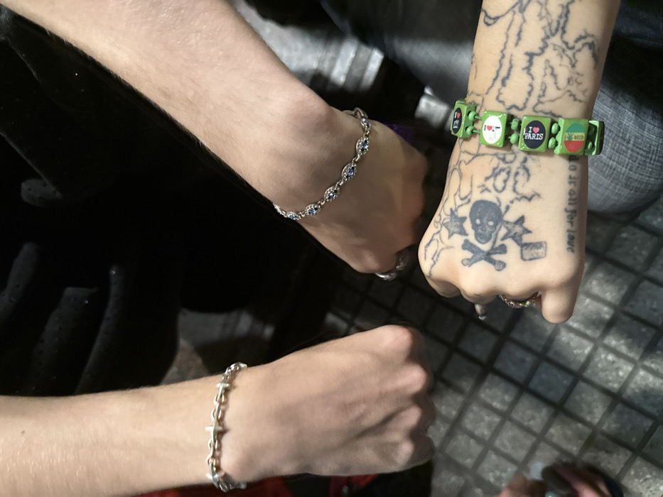 Three wrists are visible in the frame. One wears a silver bracelet with blue stones. Another has a green bracelet with square charms, including "I LOVE PARIS." The hand is tattooed with a skull and crossbones, stars, and the phrase "Do it all for love." The third wrist is adorned with a silver chain bracelet. The hands are in a fist position. The background includes dark fabric and a tiled surface.