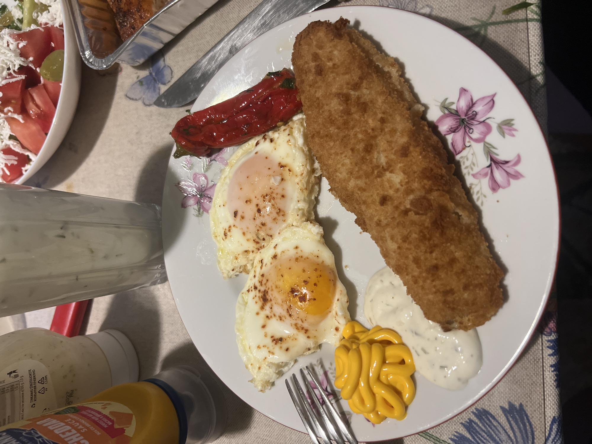 An overhead indoor shot captures a meal served on a patterned tablecloth featuring butterflies and flowers. The main subject is a white plate holding a breaded, fried item (possibly fish or schnitzel), two fried eggs with runny yolks, and a roasted red pepper. Two sauces are squirted onto the plate: a white, possibly tartar or yogurt-based sauce with herbs, and a yellow, likely cheese or mustard sauce. A metal fork rests on the plate.
Adjacent to the plate, a white bowl filled with salad, including diced tomatoes and grated white cheese, is partially visible. Further back, a metal serving tray with a cooked item is also partly seen. A clear glass containing a milky liquid stands nearby.
In the foreground, two bottles of sauce are visible: a yellow bottle labeled "CHEDDAR SAUCE" with additional text "SOS CU BRANA," "PERFECT," and nutritional information in Bulgarian ("мега 3 мастни киселики в /100 g 2.0 g"), along with an expiration year "2027". A white bottle next to it shows recycling information, including "TINKAMAI IŠRŪŠIUOKITE" (Lithuanian for "Sort correctly") and "ИЗХВЪРЛЯЙ ПРАВИЛНО" (Bulgarian for "Dispose properly"). The presence of these languages, combined with the location context, places the scene in Yardzhilovtsi, Bulgaria. The lighting suggests an evening meal.