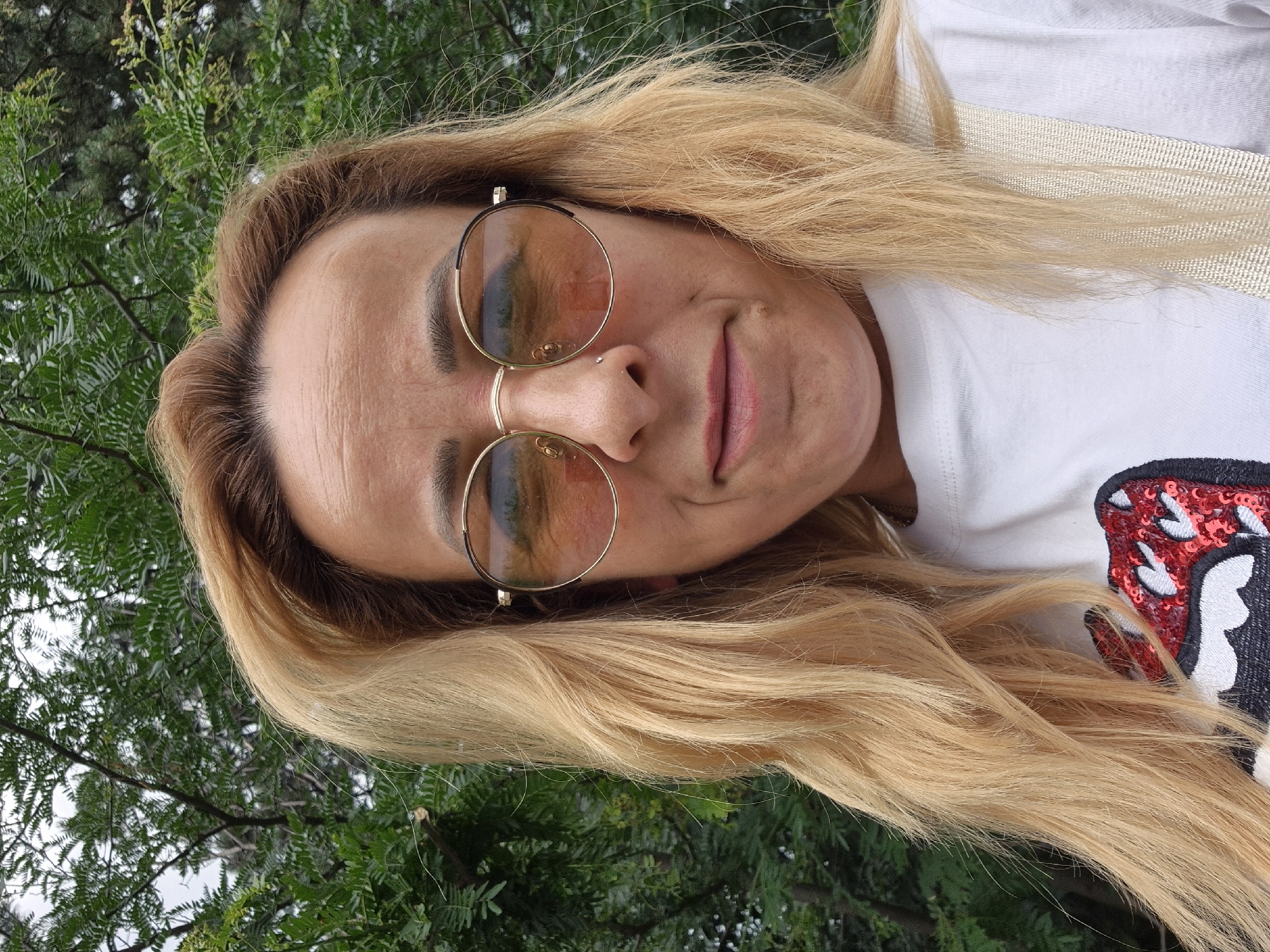 A medium shot portrait of a woman with blonde hair and sunglasses is presented against a backdrop of green foliage. The woman is wearing a white t-shirt with a red and black sequined design on the front. She has a nose ring and appears to be looking slightly off-camera. The environment suggests an outdoor setting, possibly a park or garden.