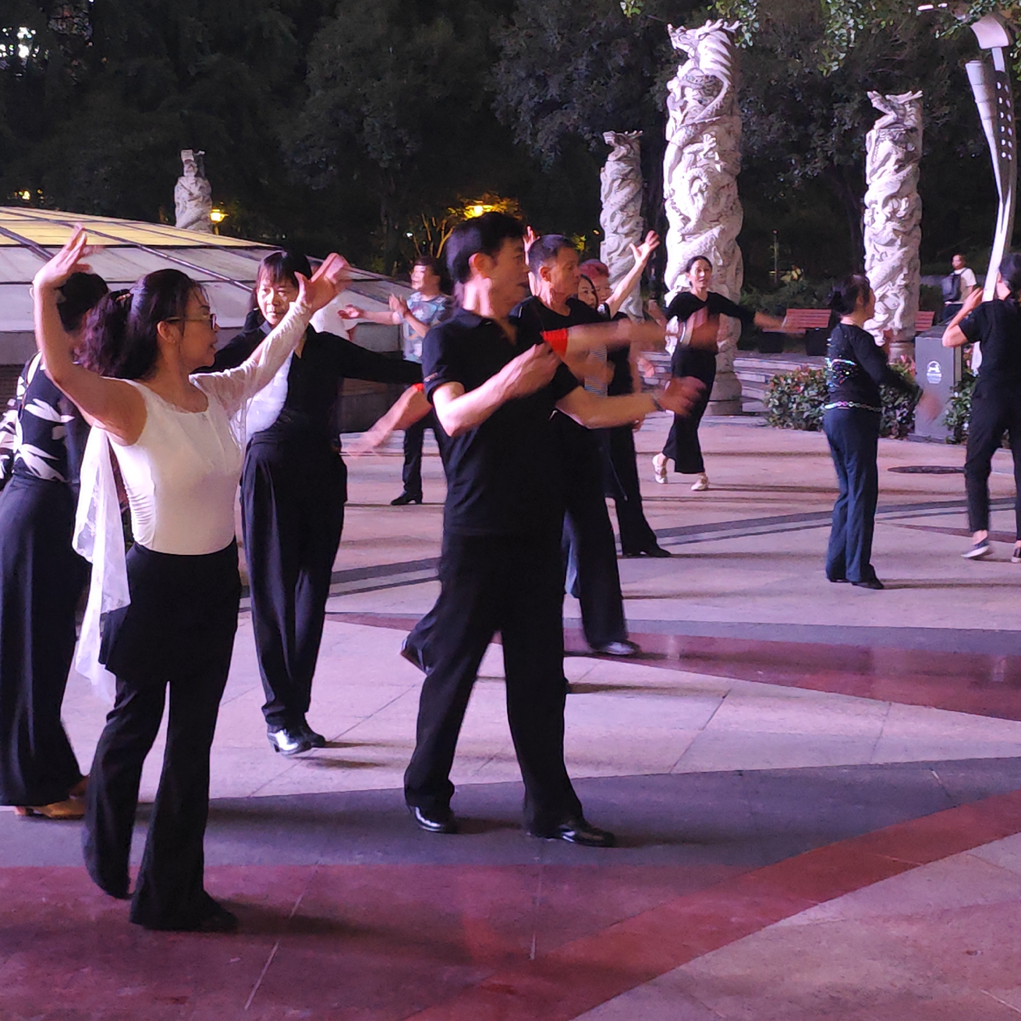 A group of people are engaged in a dance activity outdoors in the city of Liangjiang, China, at night. The participants appear to be mostly middle-aged and elderly adults. They are wearing dance attire that predominantly consists of black clothing, with some individuals having white tops. The dancers are positioned on a paved area marked with red and white lines. The dancers are in mid-movement, with arms outstretched. In the background, there are ornamental columns sculpted with dragons, a structure with a roof, trees, benches, and street lighting. The scene is illuminated artificially, creating a pinkish hue on the ground.