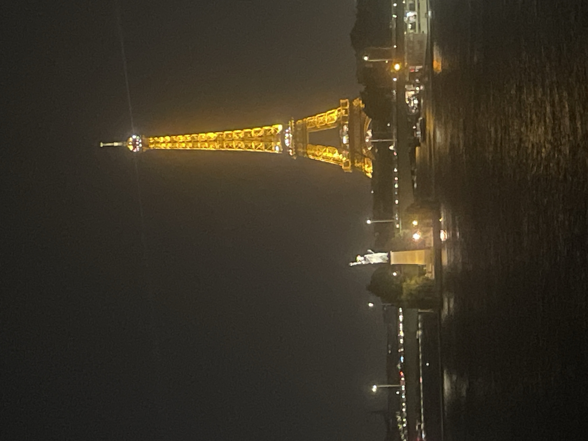 The image depicts a nighttime view of the Eiffel Tower in Paris, France, illuminated with golden lights. In the foreground, a dark body of water, likely the Seine River, reflects the distant lights. Across the river, a bridge is visible with cars and streetlights. To the left of the Eiffel Tower, a statue resembling the Statue of Liberty stands on a pedestal. The sky is dark, with no visible stars or moon. The overall scene is serene and iconic of Paris at night.