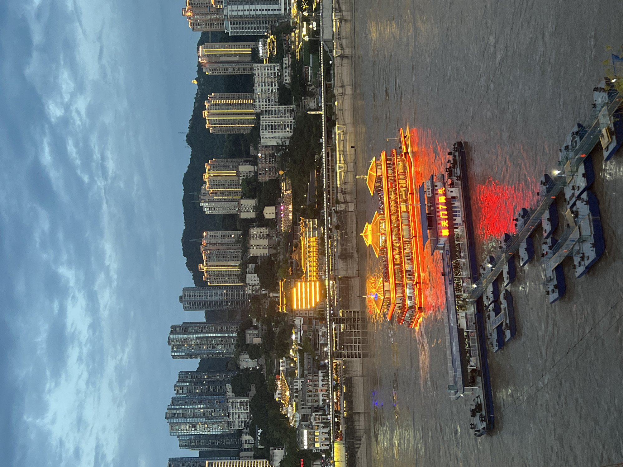 The image depicts a wide, murky river at dusk in Yuzhong District, China. Two large, illuminated tour boats are prominent on the water. The larger boat, nearer to the background, is elaborately decorated with red and gold lights, highlighting its multiple decks and people gathered on them. A smaller, blue and white boat is docked closer to the foreground, also with people visible on its deck. Connected to this smaller boat is a series of floating docks, some of which also have people present. The riverbanks are lined with a dense collection of modern high-rise buildings, many of which are illuminated with warm yellow lights. The buildings rise steeply up a hill, with some older, ornate structures visible at a lower level closer to the river. The sky is overcast with dramatic clouds, suggesting the transition from day to night. The overall scene conveys a vibrant, urban waterfront at twilight.