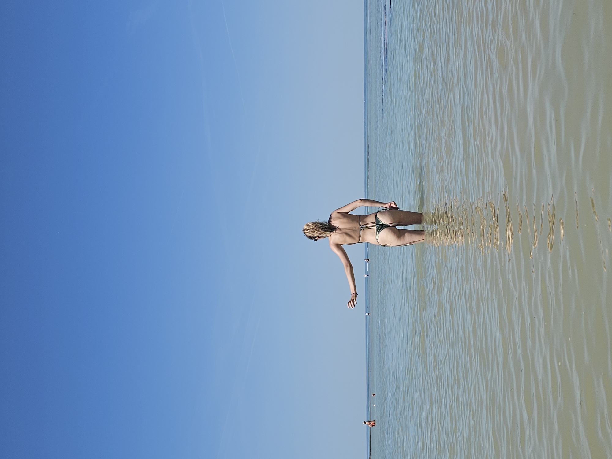 A woman in a bikini stands in shallow water, extending one arm. The location is the coast of La Brée-les-Bains, France. The sky is clear and blue. The water surface is rippled. Other people are visible further out in the water.