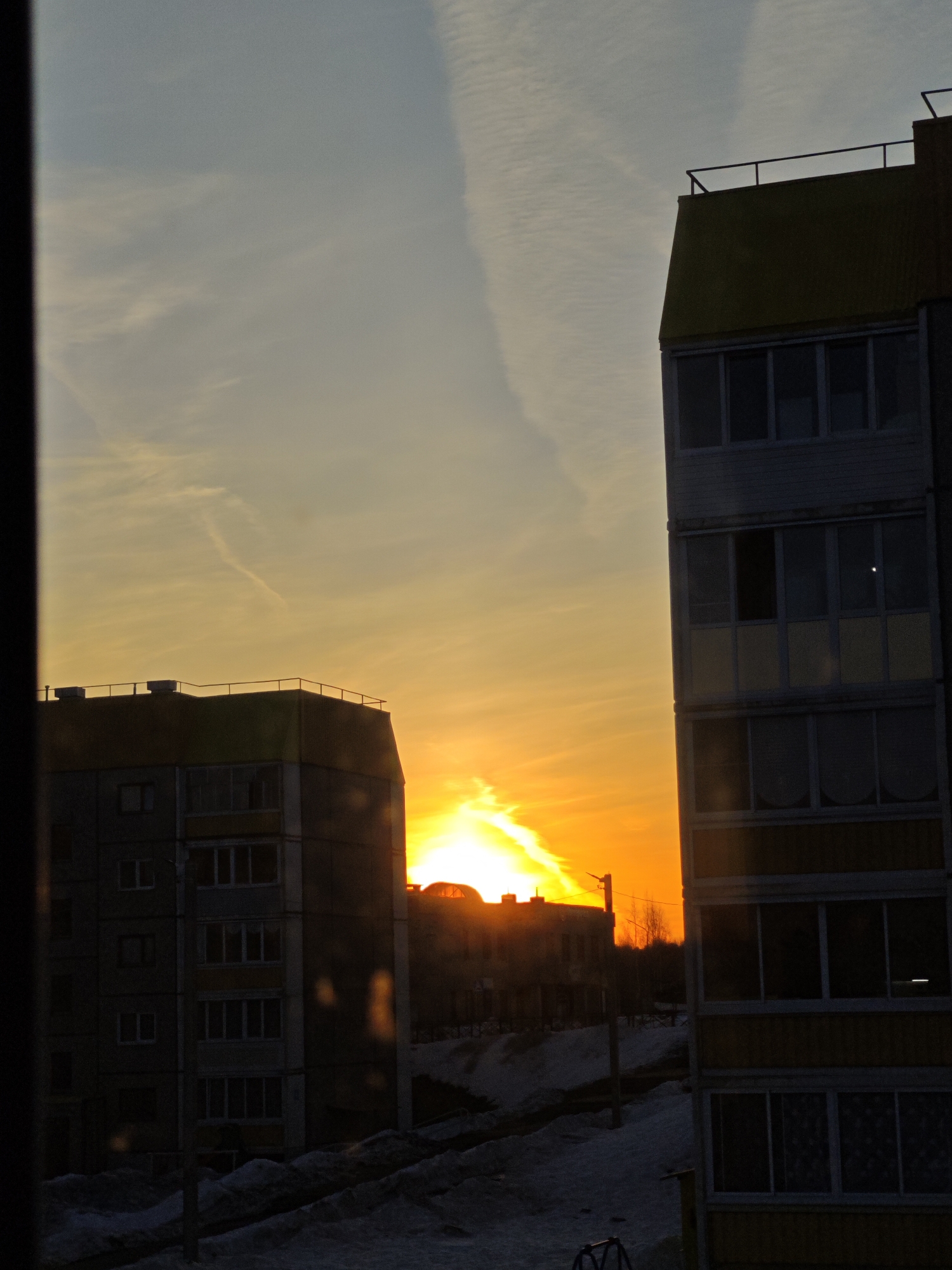 The image captures a vivid sunset viewed from within a building, likely an apartment, in Марковский, Russia. The scene is framed by the interior of the window and shows parts of two multi-story residential buildings on either side, silhouetted against the brightly lit sky. The setting sun emits a strong orange glow, partially obscured by the buildings. The sky exhibits hues of yellow and orange, with contrails visible above. Below, a snow-covered landscape can be seen, suggesting winter. Additional buildings and trees are silhouetted in the middle distance. Utility poles and bicycle are also visible.