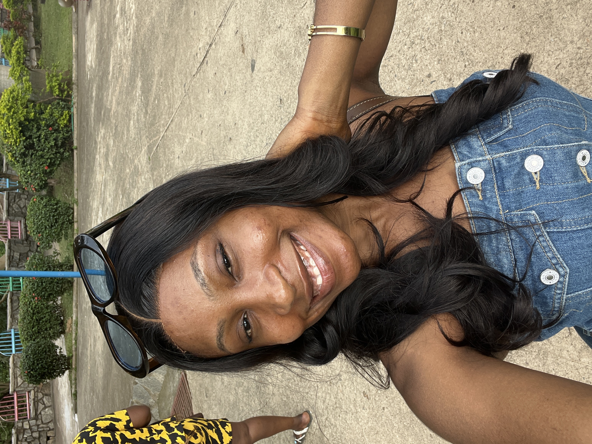 The image is a selfie of a young Black woman taken outdoors during the day. She is wearing a denim dress and a yellow and black patterned garment is visible at the bottom of the frame. She has long, dark, wavy hair and is wearing sunglasses perched on her head. A gold bracelet is visible on her left wrist. She is smiling broadly, showing her teeth, and appears happy and relaxed. The background suggests an outdoor setting with a concrete ground, some greenery and trees, and what appears to be a playground structure with colorful bars in the distance. The lighting indicates it is daytime. The overall impression is that of a casual, joyful moment captured outdoors.