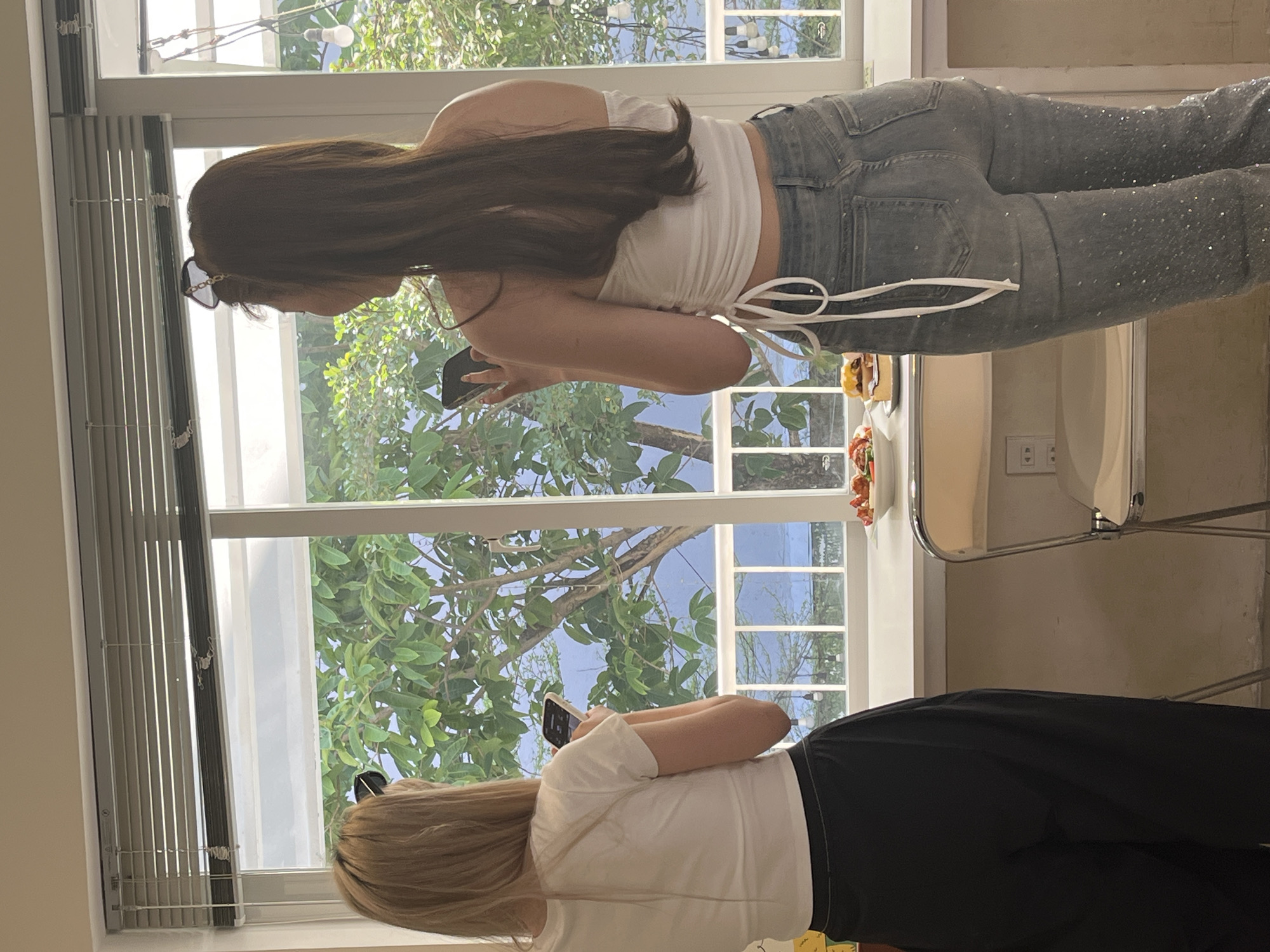 Two women are standing by a window, each looking at their phone. The woman on the left has long brown hair and is wearing a white top and jeans. The woman on the right has blonde hair and is wearing a white top and black pants. The window looks out onto a green leafy tree. There are blinds on the left side of the window. The location could possibly be in Đà Nẵng, Vietnam, given the foliage and architectural style but this is unconfirmed. There is a small table with some food on it near the window. There is a chair next to the table.