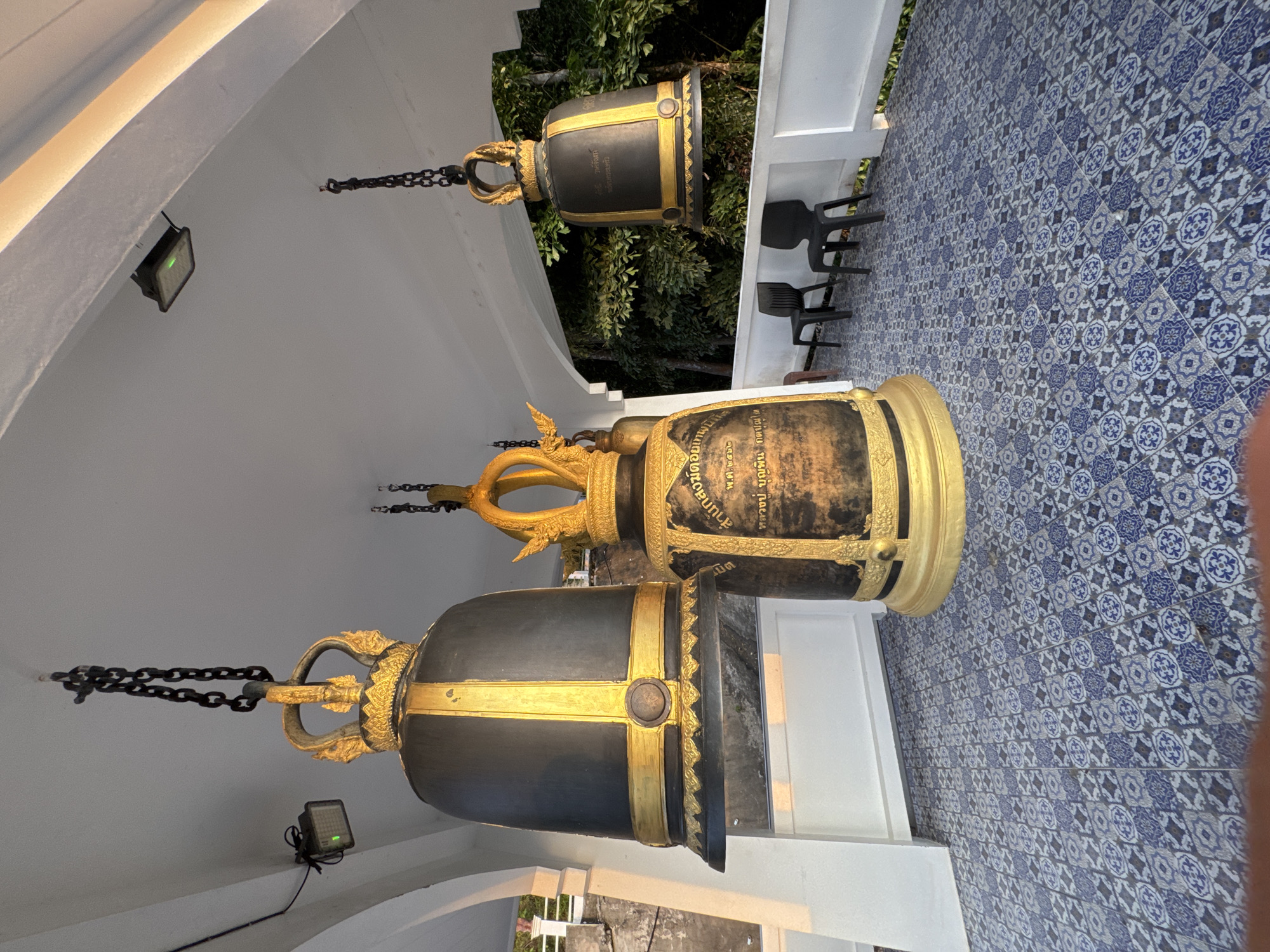 The image depicts several large, ornate bells hanging from chains under a white ceiling with recessed lighting. The bells are black with gold decorative bands and crowns. The floor is tiled with a blue and white pattern. In the background, there are two black plastic chairs and lush green trees. The scene is likely located outdoors, possibly on a balcony or covered walkway. The overall impression is of a temple or a place of worship. The presence of the bells, the architecture, and the surrounding vegetation suggest a location in Southeast Asia. While the specific location within Patong, Thailand, cannot be definitively determined from the image alone, it is a plausible setting for such a scene.