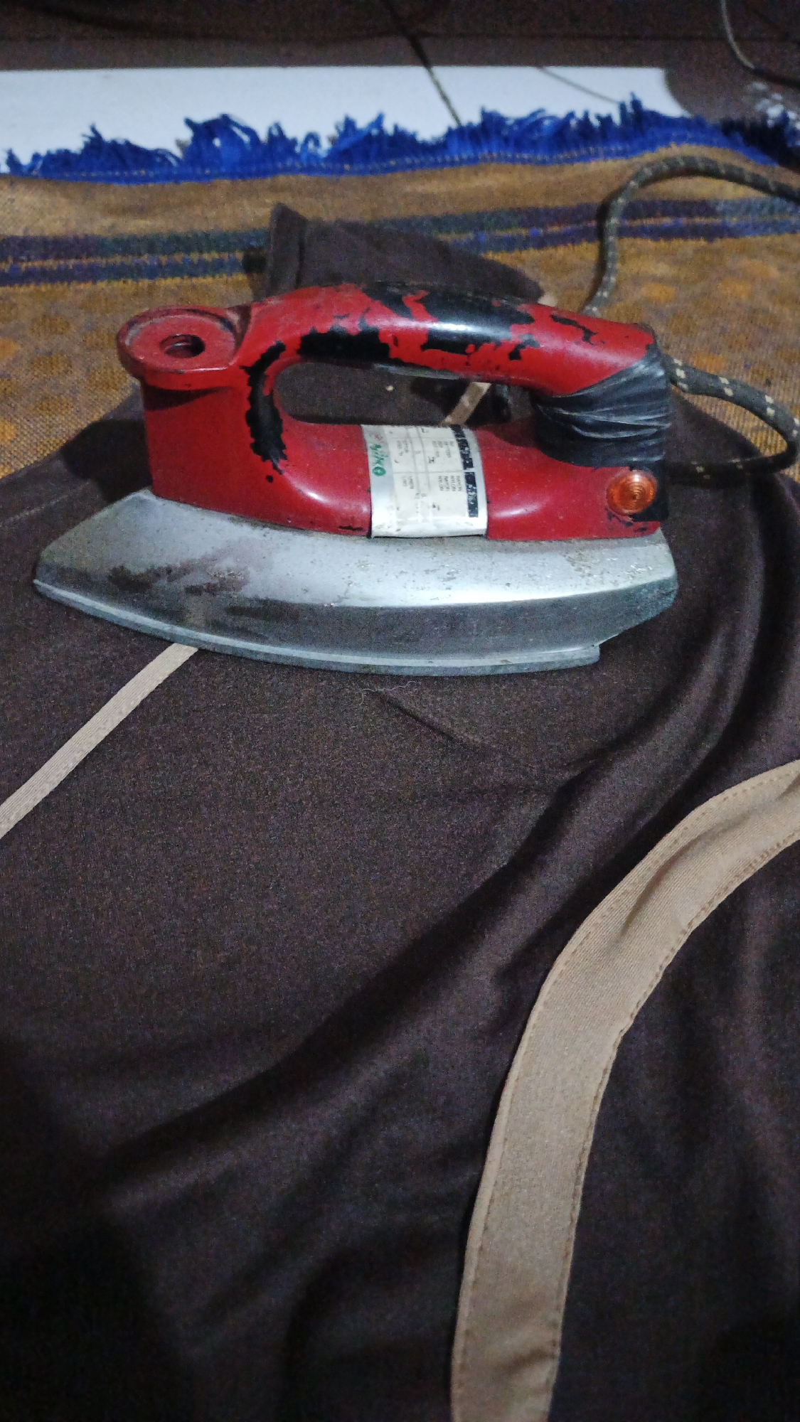 An old, red and black electric iron rests on a dark brown fabric. The iron has a worn appearance with chipped paint on its handle. A white sticker with text and a circular orange indicator light are visible on the iron. The fabric it is on is wrinkled and appears to be part of clothing or upholstery, with a light brown trim detail running across it. In the background, a striped rug with blue fringed detailing is visible. The overall impression is one of an indoor setting, likely a home, in Gunungreja, Indonesia.