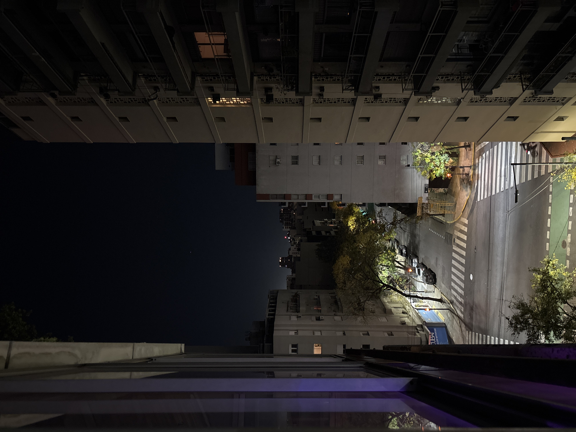 At night, a high-angle view from what appears to be an apartment building looks down onto a street in Buenos Aires, Argentina. The buildings on either side of the street are lit with some windows showing light, and the dark night sky is visible above. The street below is illuminated by streetlights, revealing a crosswalk and road markings. Some cars are parked along the side of the street. Trees are visible on both sides of the road, their branches illuminated by the streetlights. The perspective is unusual, with the top of the image showing a darker scene than the bottom. The viewer seems to be looking out through a window, with part of the window frame visible in the foreground.