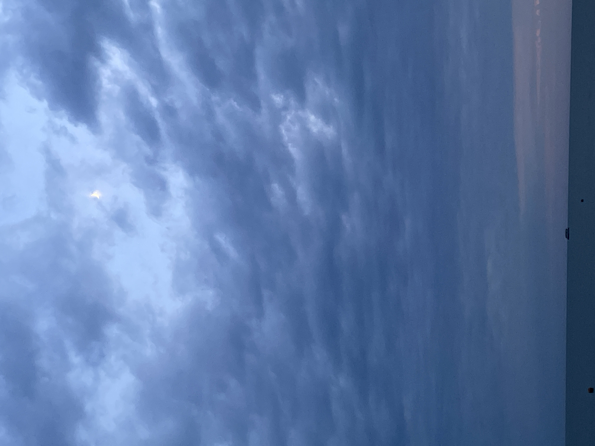 The image shows a dark, cloudy sky above a body of water. A sliver of the moon is visible in the upper right quadrant of the sky, appearing as a pale crescent. The clouds are dense and textured, with varying shades of blue and gray. Towards the horizon, the sky transitions to a lighter, pinkish hue. The water is calm and reflects the muted colors of the sky. In the distance, a small boat is visible on the water. There are no people or specific landmarks visible in the image. The scene suggests an evening or early morning setting, possibly near a coast or large lake.