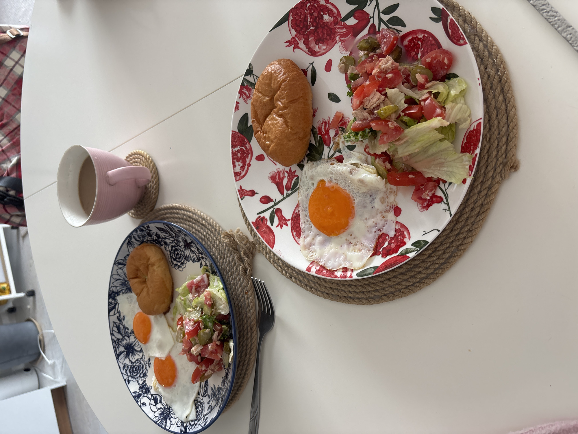 Two plates of breakfast are laid out on a white table. Each plate contains two fried eggs, a small bun, and a salad made with chopped tomatoes, lettuce, and what appears to be tuna or chicken. One plate has a blue and white floral pattern, while the other features a red pomegranate design. A pink mug of coffee sits on a coaster to the right of the plates. A fork is visible on the table between the two plates. The background shows a glimpse of a kitchen or dining area with a white cabinet and a partially visible patterned blanket. There is no information available to determine the location of this scene within Krasnoyarsk, Russia.