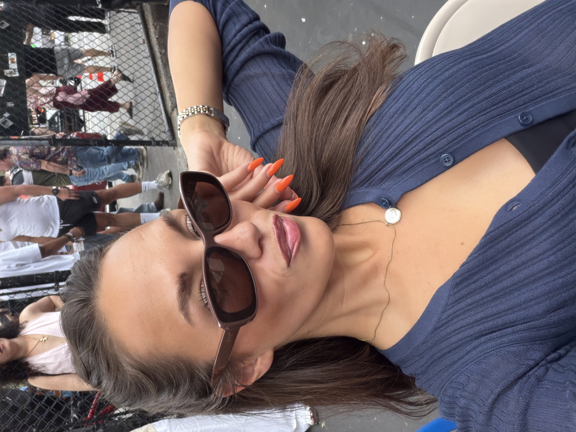 An outdoor, eye-level photograph captures a woman with dark brown hair and a tanned complexion, wearing dark brown rectangular sunglasses. Her lips are glossy with a reddish-plum tint and are slightly parted. She is dressed in a navy blue ribbed knit V-neck cardigan, unbuttoned over a black undershirt, and wears a thin gold chain necklace with a circular silver pendant. Her left hand, featuring long, pointed nails painted bright orange, is positioned near her right temple. A silver and gold-toned link bracelet is visible on her left wrist.
The background, seen through a dark chain-link fence, depicts a bustling urban street scene under daylight. Numerous individuals in casual attire, including t-shirts, shorts, and jeans, are present. Notable figures include a woman in a light pink halter top with a gold cross necklace visible in the upper left, and a man in a white t-shirt and dark shorts to the left. A dark vehicle and at least one orange traffic cone are partially visible through the fence, indicating a pedestrian area adjacent to a roadway. The ground appears to be dark pavement. The scene is located in New York City, United States of America.