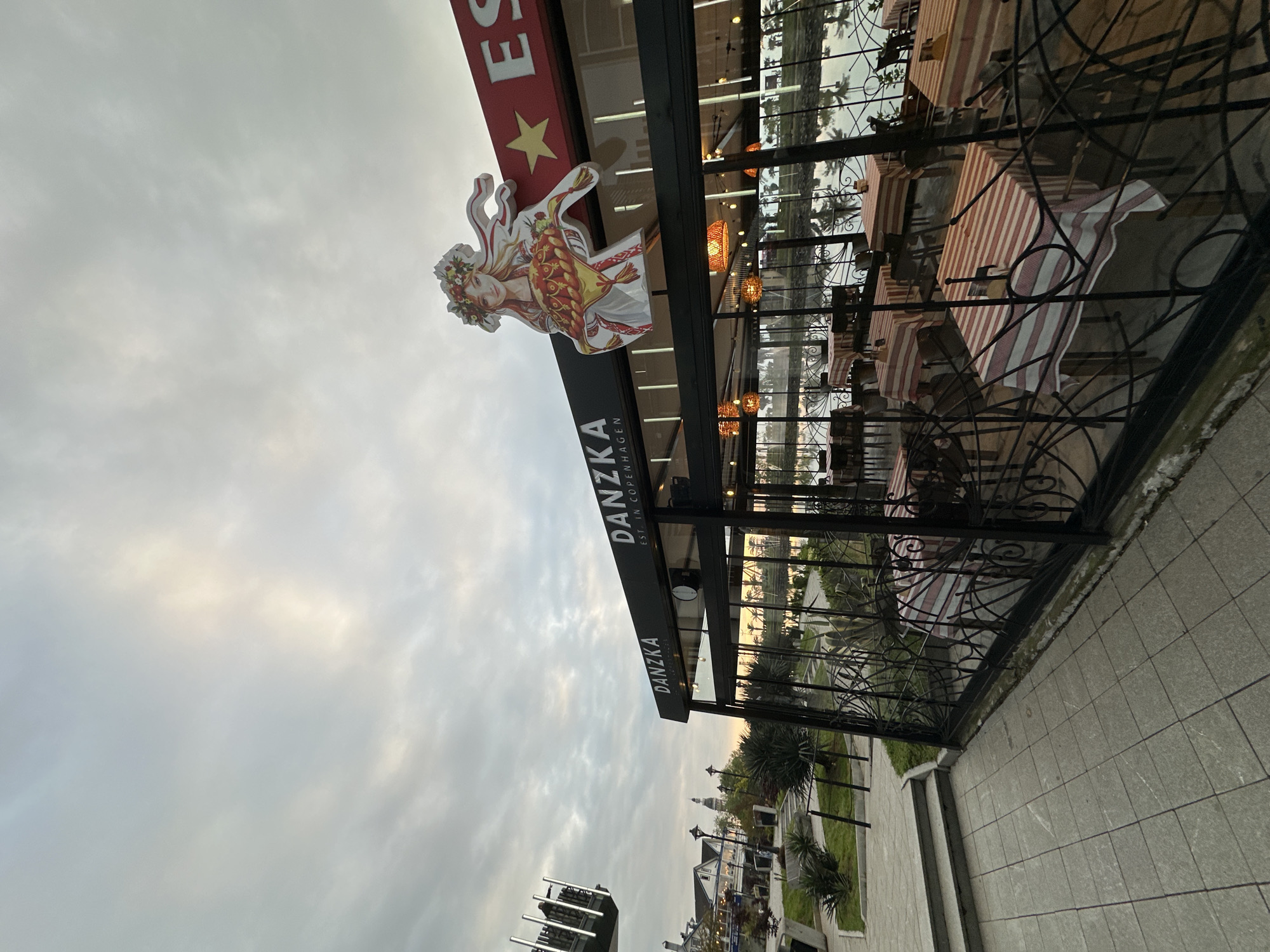 The image shows an outdoor dining area of the "Danzka" restaurant in Batumi, Georgia. The restaurant's sign is visible, featuring the brand name and a stylized image of a woman in traditional attire. The dining area is enclosed by a black iron fence. Tables with red and white striped tablecloths are arranged within the enclosure. The sky is overcast. In the background, across a body of water, buildings and vegetation are visible. A paved walkway runs alongside the dining area. Street lights and additional buildings are visible further down the walkway.