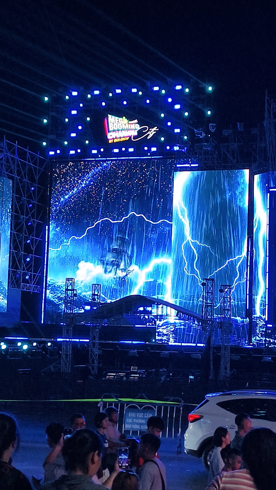 An outdoor nighttime event is underway in Nha Trang Ward, Vietnam, featuring a large LED screen displaying a dramatic scene of a sailing ship amidst a stormy sea with lightning strikes. The screen is framed by scaffolding and stage lighting, with blue and green lights illuminating the structure. In the foreground, a crowd of people are gathered, many holding up their phones to capture the event. A white SUV is parked to the right, partially obscured by the crowd. A sign with Vietnamese text, likely indicating restricted areas, is visible behind a fence in the mid-ground. The atmosphere appears festive and energetic.