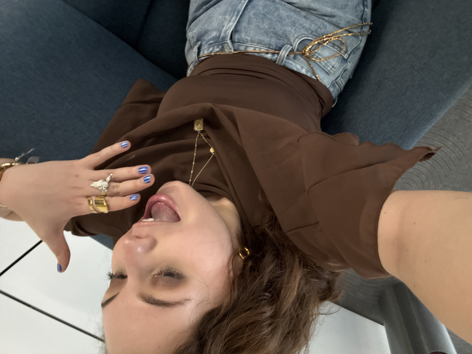 A young woman with brown hair is lying on a blue couch, her head tilted back, mouth open, and tongue visible. She is wearing a brown top, light blue jeans with a gold chain belt, and various pieces of jewelry including rings on her fingers, a bracelet on her wrist, earrings, and a necklace. Her fingernails are painted blue. The background is a gray floor. The location is unknown.