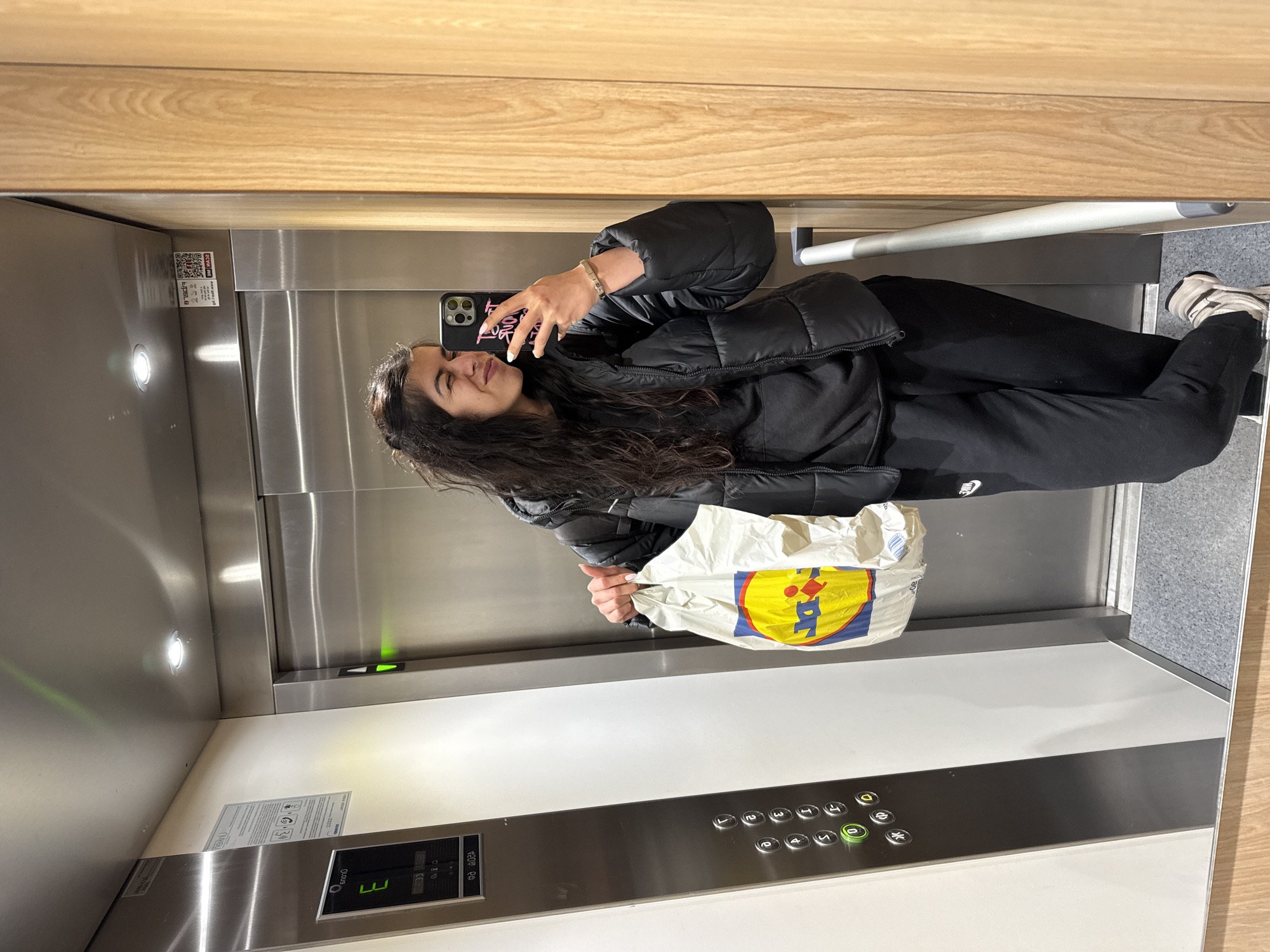 A young woman is taking a selfie in an elevator. She's holding a shopping bag with the Lidl logo. The elevator has a control panel with buttons, including one lit up for floor 0. The elevator interior is stainless steel and white. The woman is wearing a black puffer jacket and black pants. The environment suggests an urban setting, and the presence of Lidl indicates that the location is potentially Sofia, Bulgaria, as Lidl is a common supermarket chain in the city.