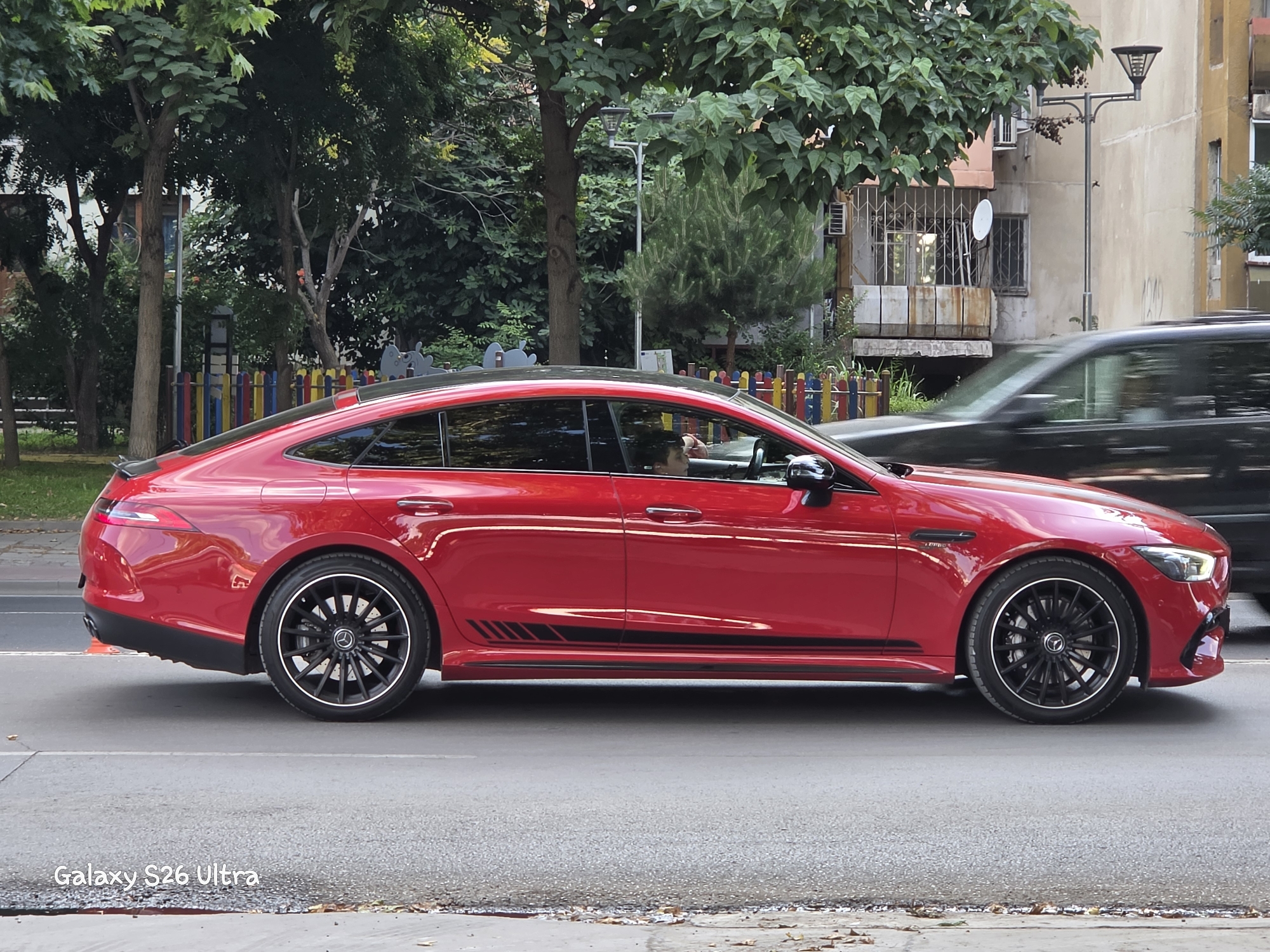 A bright red Mercedes-AMG GT 4-Door Coupe is captured in a side profile shot, driving on a paved road. The car features black racing stripes along its lower body and black rims with red brake calipers. A person is visible inside the car, seated in the driver's seat.
The setting appears to be urban, with trees and buildings in the background. A colorful fence, possibly part of a playground, is visible behind the car. A street lamp stands to the right of the car, and other vehicles, including a blurred black car in the foreground, are also present, suggesting traffic.
The lighting suggests it is daytime, with natural light illuminating the scene. The weather appears to be overcast or the lighting is muted, as there are no strong shadows. The overall impression is a snapshot of a car in motion within a European city environment. The text "Galaxy S26 Ultra" is watermarked at the bottom left of the image.