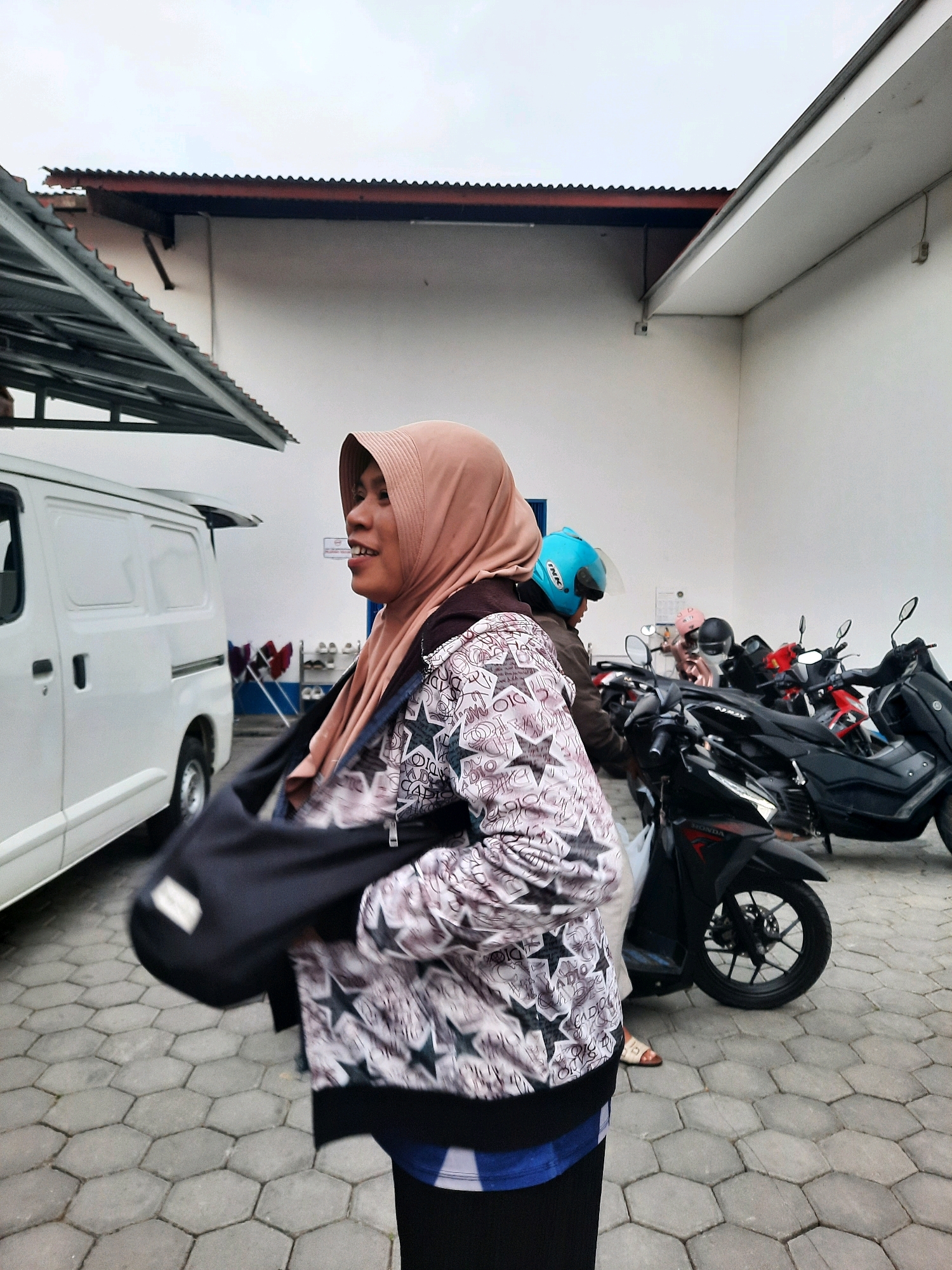 A woman wearing a light pink hijab and a patterned jacket walks in front of a white van and several parked motorcycles in Andong, Indonesia. The woman has her left arm across her chest, holding a dark bag. Her mouth is open as if she is speaking or laughing. The ground is paved with hexagonal gray tiles. In the background, there are buildings with white walls and corrugated metal roofs. The sky is overcast.