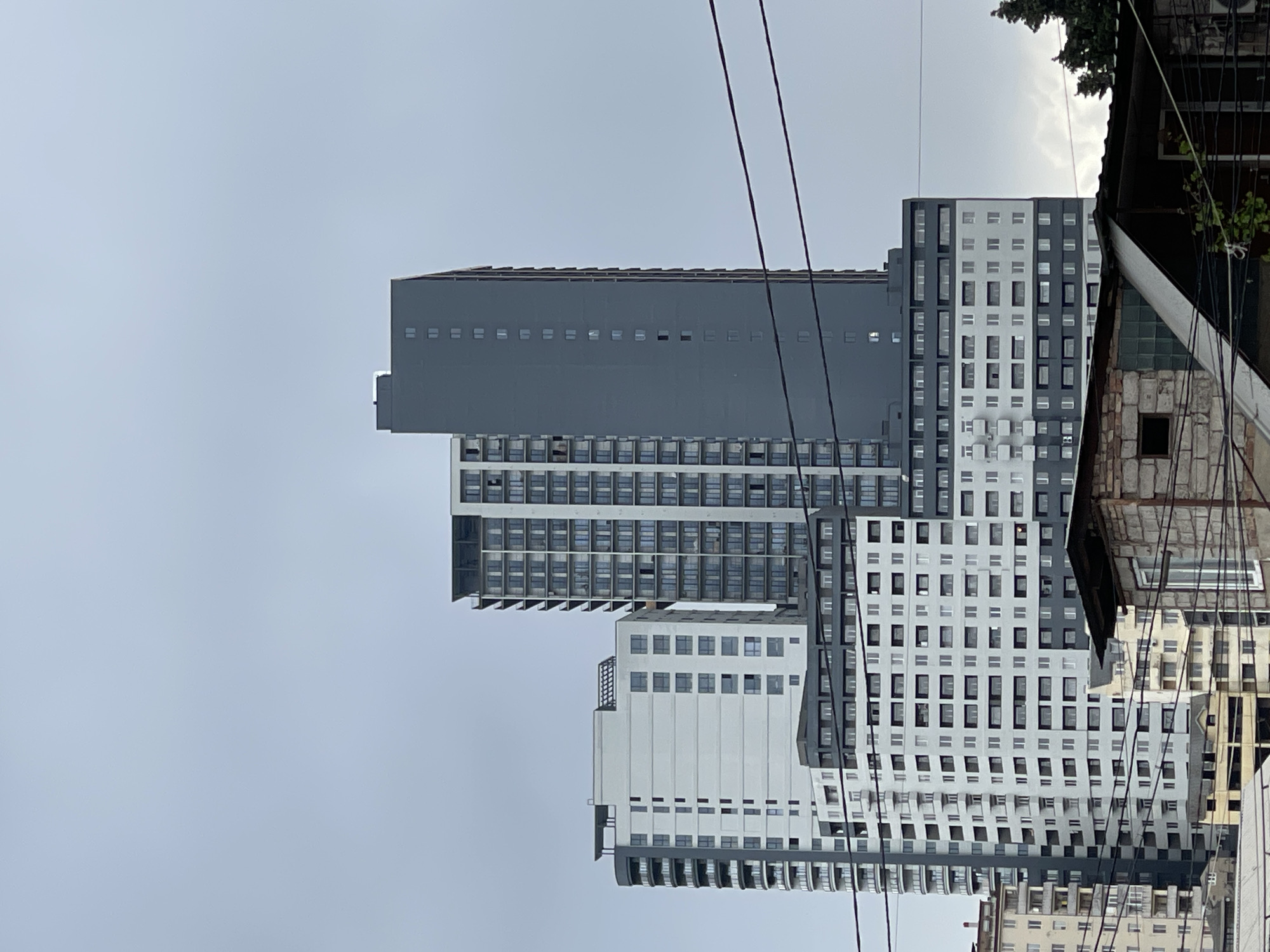 A medium, slightly rotated shot showcases modern, high-rise apartment buildings in Tbilisi, Georgia, under an overcast sky. The architecture is a mix of gray and white structures with multiple rows of windows. Power lines run across the sky in the foreground. The right side of the image features an older building, partially dilapidated, with trees visible at the top. The environment suggests an urban setting undergoing development.