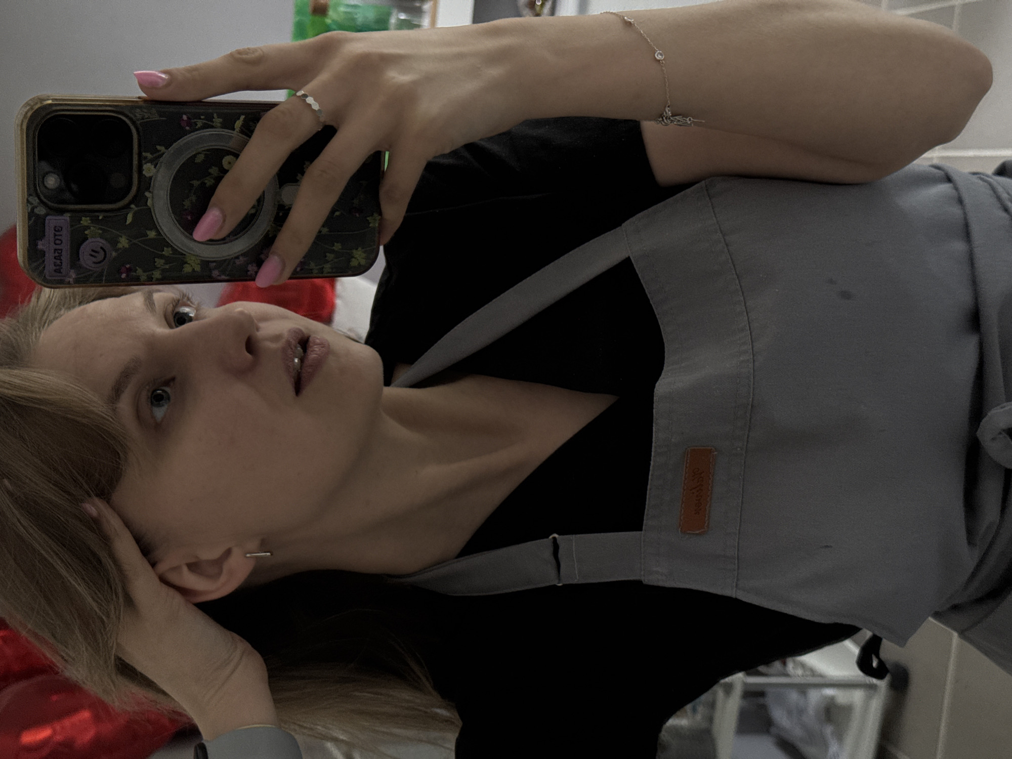 A person is taking a selfie. The person has blonde hair and is wearing a black shirt with a grey apron. They are holding a smartphone with a floral-patterned case in their hand, and the screen is facing the camera. The person is also wearing a ring and a bracelet. The location cannot be determined from the image.