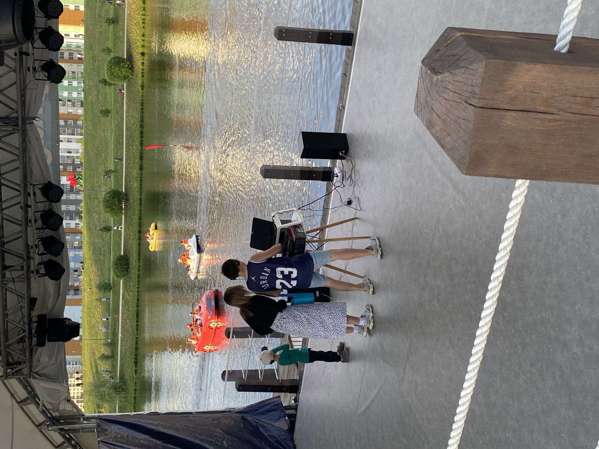 The image shows a scene from the city of Yekaterinburg, Russia, depicting a stage overlooking a body of water, possibly a pond or lake. On the stage, there are three individuals: a man wearing a "JORDAN 23" jersey, a woman in a patterned skirt, and a child in a green shirt. They are standing near some technical equipment, including speakers and what appears to be a mixing console. In the background, on the water, several pedal boats with people can be seen. Behind the water, there is a grassy area and buildings in the distance. The image is taken from a slightly elevated position, possibly from another part of the stage.