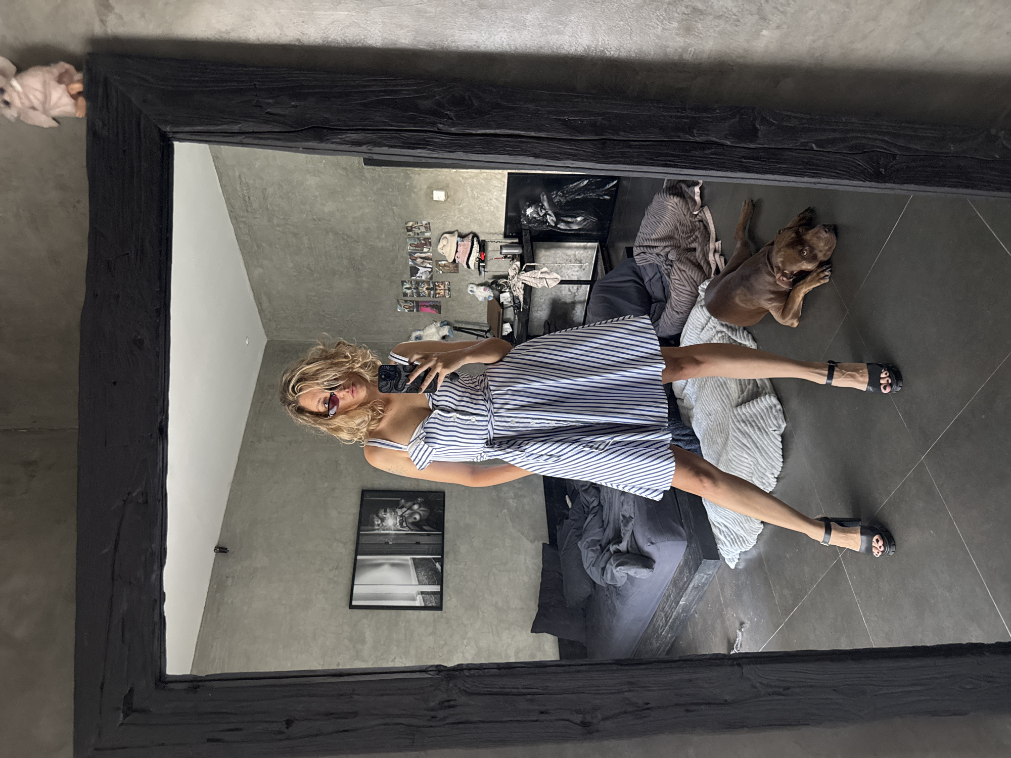 A woman is taking a mirror selfie in a room. The room has grey concrete walls and floor. The woman is wearing a striped dress and black sandals. A dog is lying on the floor next to a pile of clothes. There is a bed in the background and a black and white framed photo on the wall. There are also other photos on the wall. A stuffed animal is sitting on top of the mirror. I cannot tell if the location is in Mengwi, Indonesia.