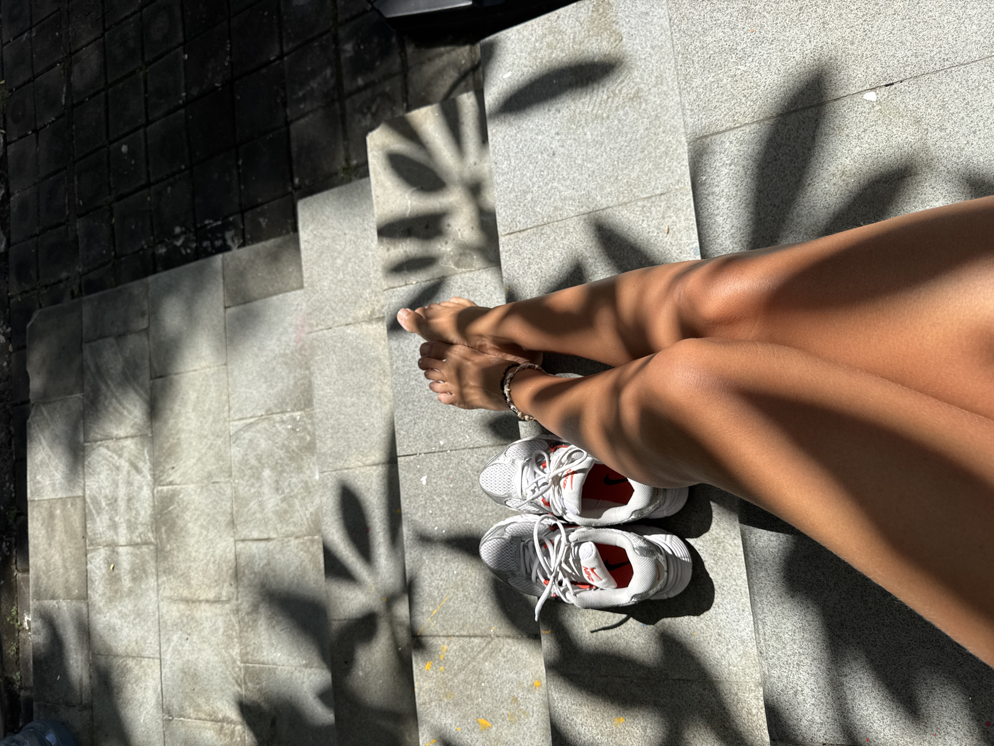 The image depicts a person sitting on concrete stairs in Kuta Selatan, Indonesia. The individual is fair-skinned with tan legs and bare feet, wearing a bracelet on their left ankle. A pair of white athletic shoes with red interiors and white laces sits on the step beneath their feet. The stairs are made of large, rectangular concrete slabs. Sunlight filters through foliage above, casting sharp shadows on the steps and the person's legs and shoes. Some of the adjacent ground is paved with dark, square tiles.