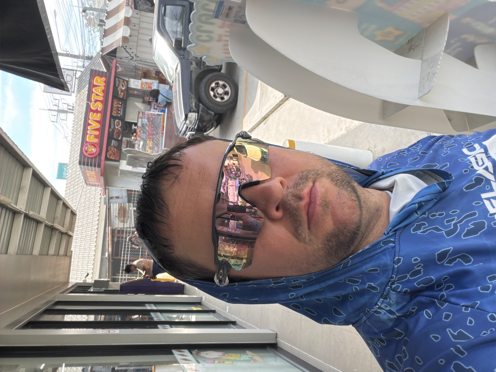 A man wearing a blue hooded shirt and sunglasses takes a selfie outdoors in Baan Mae Nam, Thailand. He is standing in front of a row of shops, with a "Five Star" fast food stall visible in the background. A dark-colored pickup truck is parked in front of the stall. The man's sunglasses reflect the street scene, showing buildings and a yellow car. To his right, a white display stand with promotional material is partially visible. The overall environment suggests a commercial area with a modest, possibly roadside, setting.