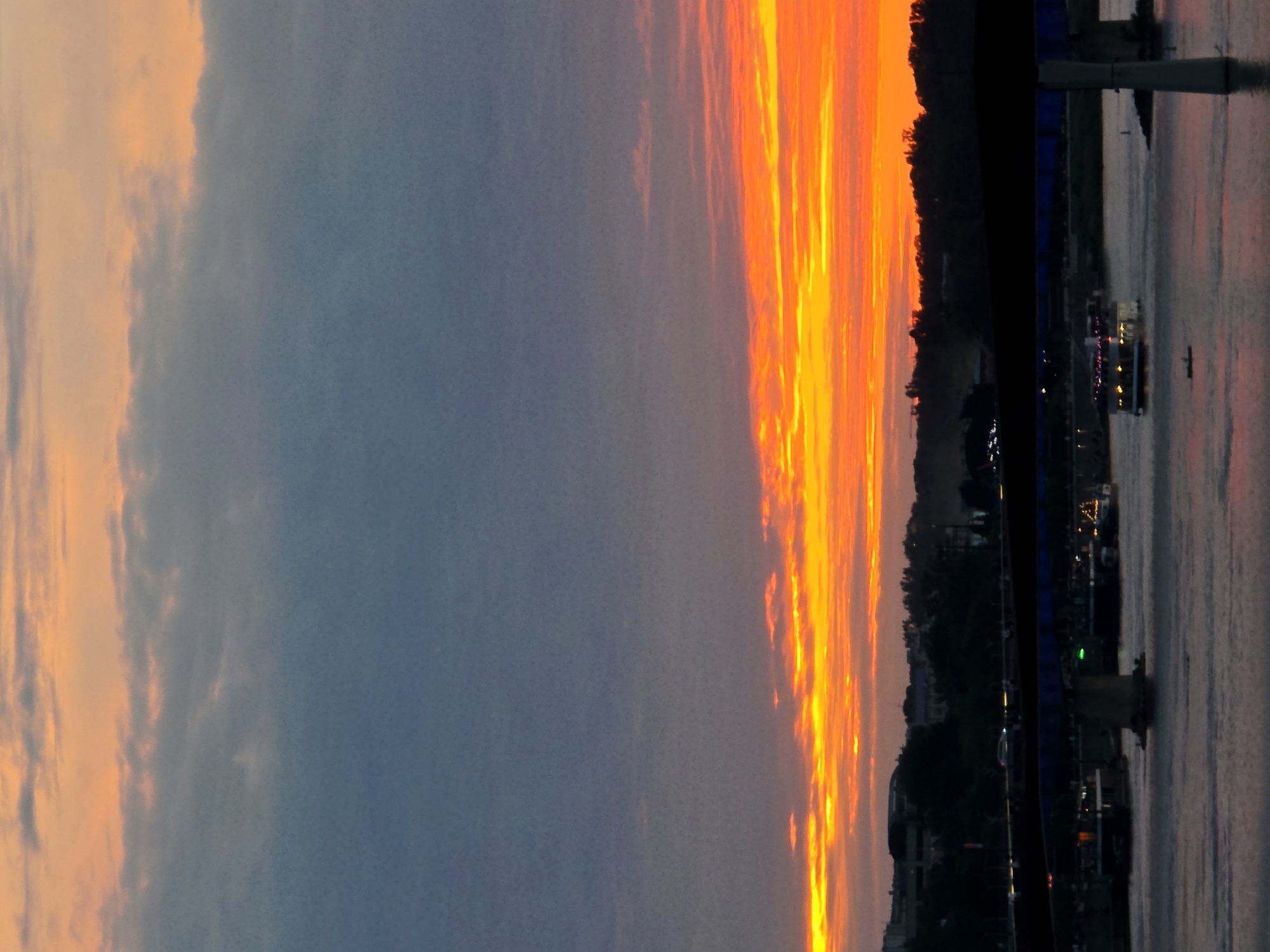 This is a photograph of a sunset over the Vistula River in Warsaw, Poland. The sky is a vibrant orange and yellow, with some grey clouds. A bridge spans the river, and below it, several boats are visible. The shoreline is lined with trees and buildings. The image appears to have been taken from a distance, possibly from another bridge or a tall building.
