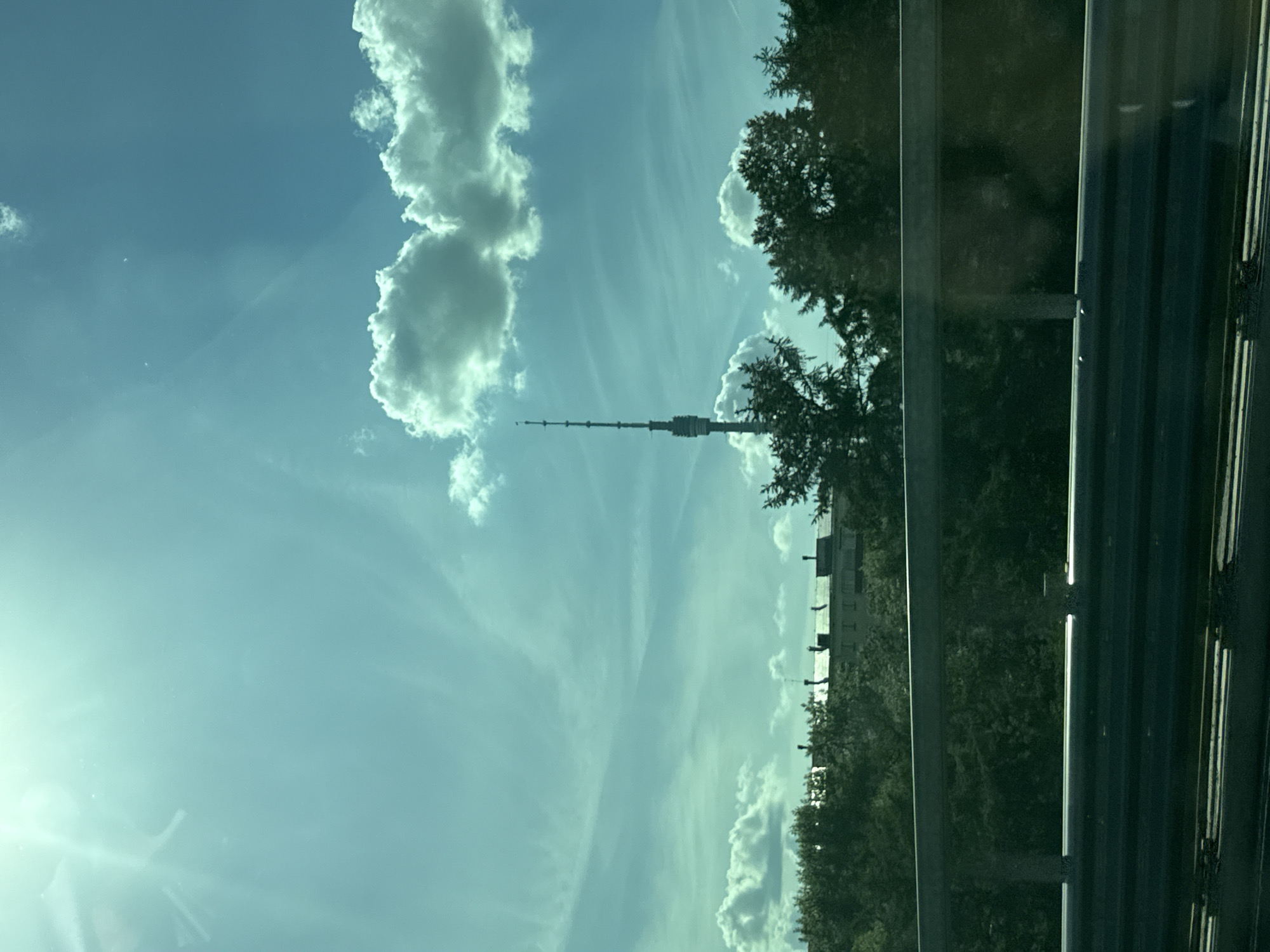 The image captures a bright outdoor scene dominated by a clear blue sky with scattered white clouds, some appearing puffy cumulus and others streaky cirrus. A powerful sun glare emanates from the upper left corner, casting a luminous glow across the sky.
In the middle ground, a tall, slender telecommunications or observation tower rises prominently towards the sky. Its structure is segmented, with the top tapering to a point. Dense green trees are visible beneath and around the tower, extending across the horizon. Beyond the trees and slightly to the right of the tower, a low-lying building or industrial structure can be seen in the distance.
The foreground on the right side of the image features dark, reflective vertical elements, which appear to be a car window frame or pillar and a metallic highway guardrail or barrier. These elements are partially obscuring the view of the trees and background. The perspective suggests the photo was taken from inside a moving vehicle, looking out at the landscape.
Given the location context of Moscow, Russia, the prominent tower is identifiable as the Ostankino Tower. The time of day is bright daylight, likely midday or early afternoon, indicated by the strong sunlight and clear skies. There is no visible text in the image.