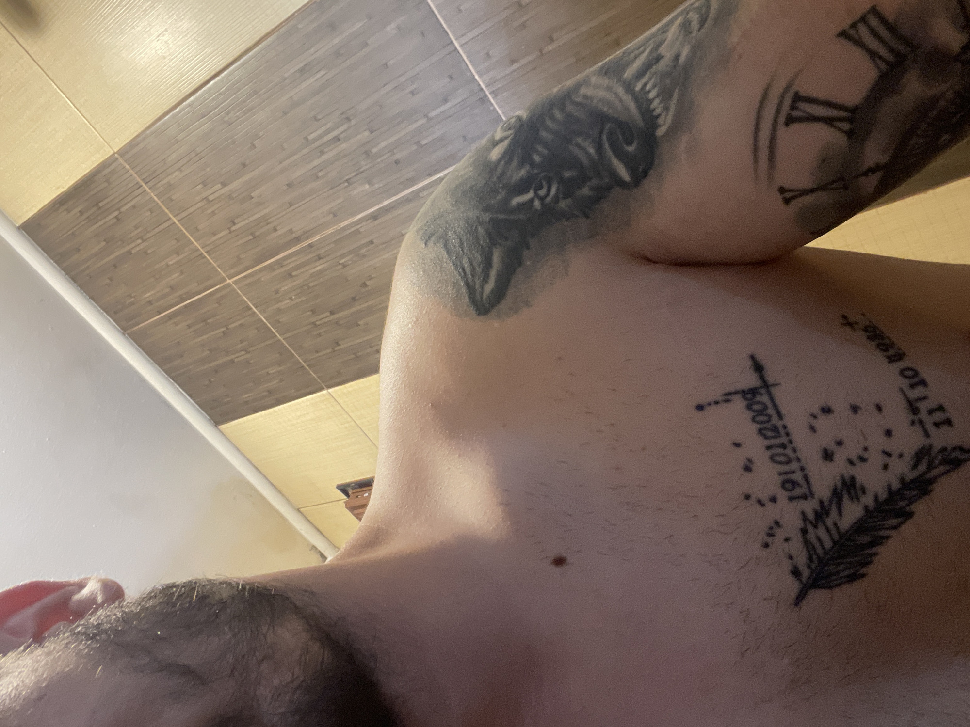 This is an indoor, close-up photograph taken from a low, upward angle, focusing on a person's upper body. The subject is likely male, evidenced by visible body hair on the chest and shoulder, and facial hair (beard or stubble) in the lower-left corner of the frame.
The person has several prominent tattoos. On their left upper arm, there is a detailed tattoo of an animal head, possibly a wolf or a bear. Further down the same arm, another tattoo depicts Roman numerals, including "X," "XI," and "II," suggesting a clock face design. On the upper right side of the chest (from the viewer's perspective), there is a smaller tattoo featuring a stylized feather, surrounded by scattered dots. Accompanying this chest tattoo are visible numbers "191010161" and partially discernible text above the feather that appears to include "11 10 Ho". A small mole is also visible on the person's upper chest/collarbone area.
The background consists of a tiled surface, likely a wall or ceiling. It features two distinct types of rectangular tiles: an upper section of lighter, possibly cream-colored tiles with a horizontal ridged texture, and a lower section of darker, brown-grey tiles with a linear, wood-grain like pattern, also ridged. The lighting suggests an indoor setting, possibly with artificial light, creating subtle shadows. The person appears to be posing for the photograph.