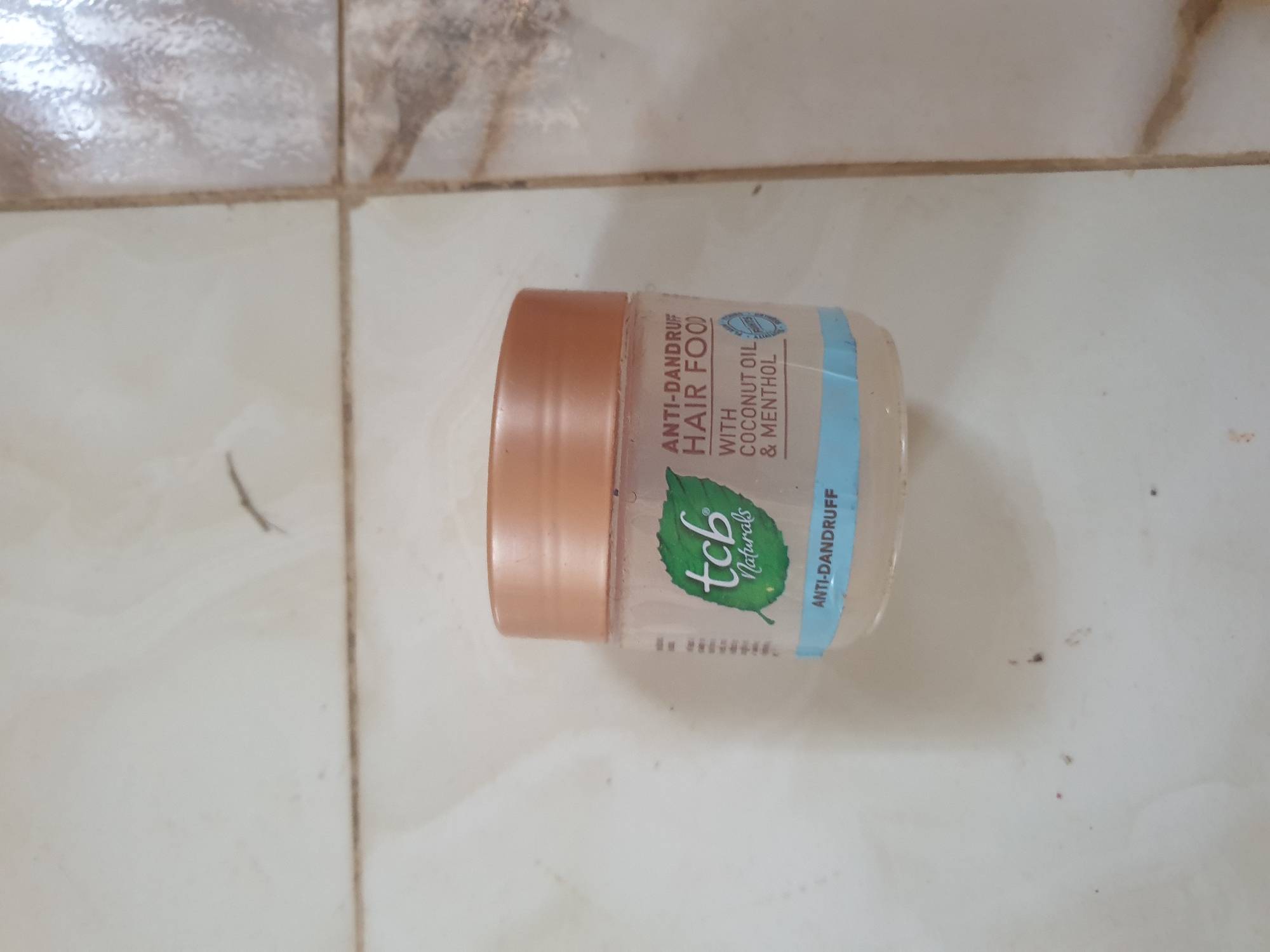 In Anilelerin, Nigeria, a small, clear plastic jar with a peach-colored lid sits on a tiled floor. The jar contains a light-colored substance and has a label with green and blue accents. The label features a green leaf graphic and text that reads "tcb Naturals," "ANTI-DANDRUFF HAIR FOOD," "WITH COCONUT OIL & MENTHOL," and "ANTI-DANDRUFF." The tiled floor appears to be light-colored with subtle patterns.