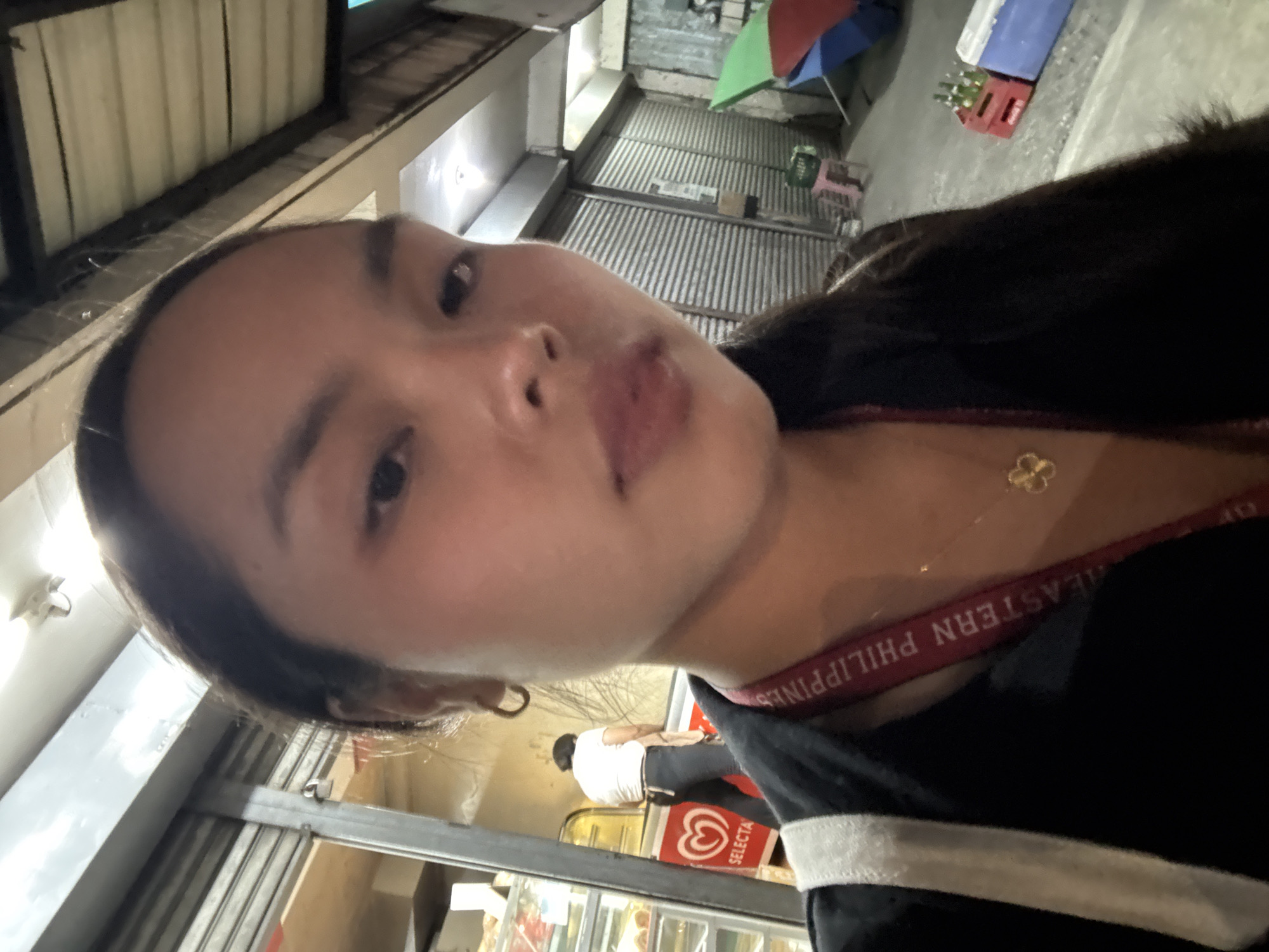 A selfie taken outdoors shows a woman with dark hair, brown eyes, and light complexion. She wears a black top with white trim and a red strap labeled "EASTERN PHILIPPINES" and a gold flower necklace. The background shows an urban environment with corrugated metal structures. In the distance are some items on the sidewalk, including a red crate, blue box, and umbrellas. A man in a white shirt is slightly visible inside a building behind the woman. A Selecta ice cream freezer is visible. The location is possibly in Davao City, Philippines, as indicated by the "EASTERN PHILIPPINES" text.